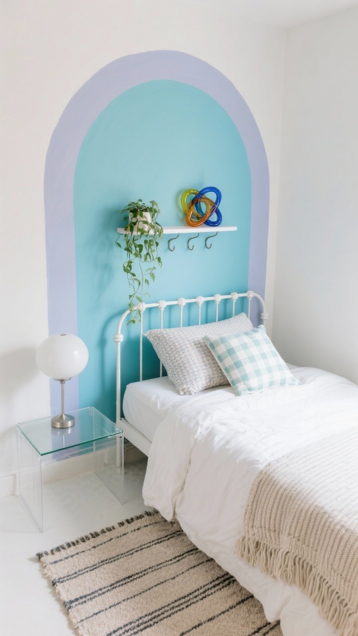 Photorealistic overhead corner angle (slight top-down) of a playful small bedroom: a soft color-blocked painted arch behind the bed in two tones—powder blue base with dusty periwinkle arch, framing the bed like a custom headboard and visually heightening the wall. Slim matte white metal bed with a crisp white duvet, patterned pillows in a tight palette including a tiny gingham accent, and a ribbed knit throw. A small glass-top nightstand keeping the footprint light; acrylic wall hooks acting as floating “nightstand” add-ons. Round globe table lamp with brushed nickel details. A petite shelf mounted inside the painted arch displaying a trailing plant and a small colorful glass knot sculpture. Flatweave rug with a thin stripe. Bright, cheerful studio-chic mood, no people.