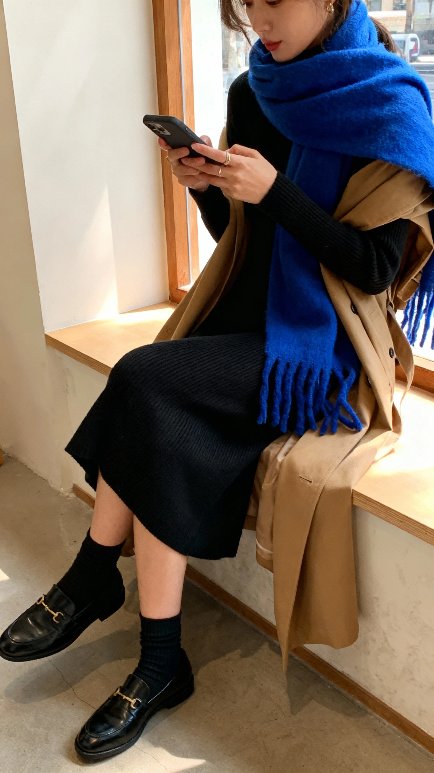 Candid cafe photo of a woman wearing a black minimalist knit dress, sleek loafers with socks, a camel trench draped over shoulders, and a bold Knicks-blue scarf for color pop, typing on her phone at a window seat, face slightly blurred, warm window light, casual iPhone aesthetic.