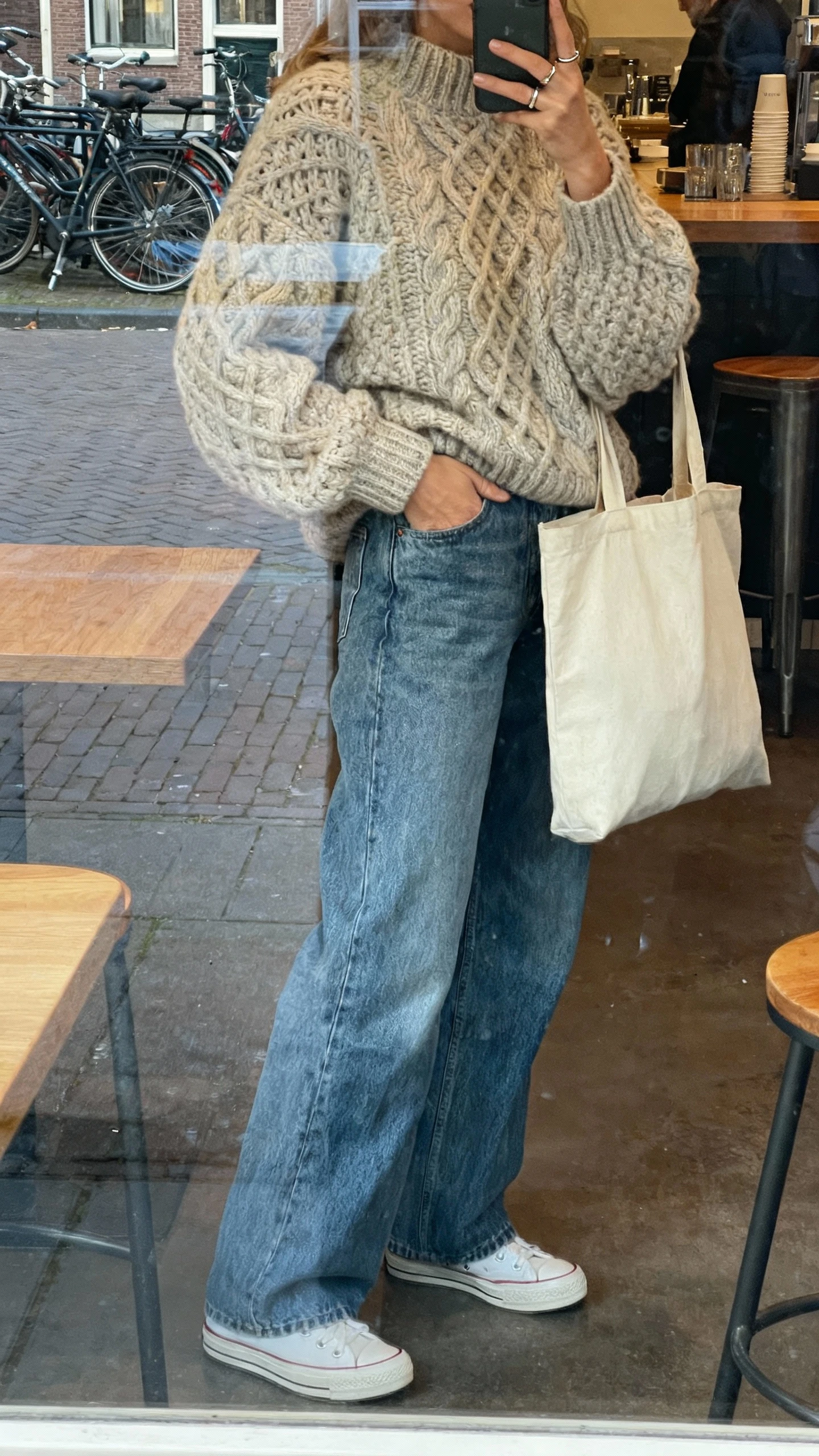 Casual iPhone selfie, side angle, of a woman in a chunky fisherman sweater, wide-leg denim, white retro sneakers, canvas tote, minimal rings; face not visible, taken in a coffee shop window reflection with bikes outside, natural daylight, Dutch-cool vibe, natural iPhone aesthetic.
