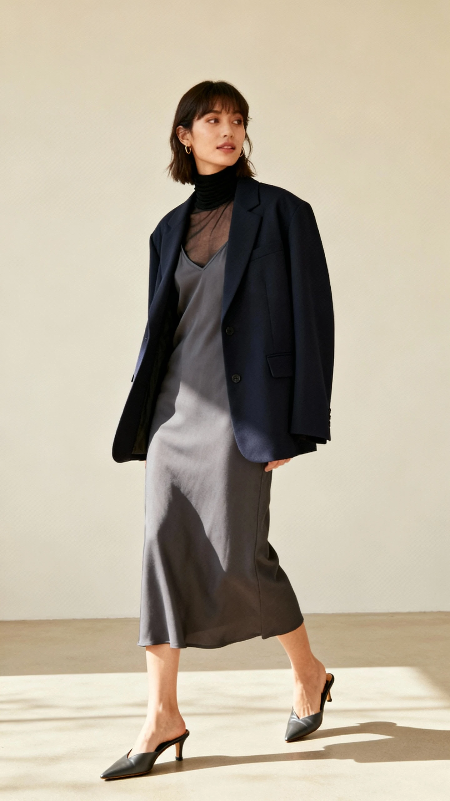 Editorial studio shot of a woman wearing a sheer black turtleneck under a layered shadow-toned look: charcoal slip dress, inky oversized blazer, and slate-length skirt, with pointed mules, cream background, studio lighting, three-quarter angle, clean and polished composition.