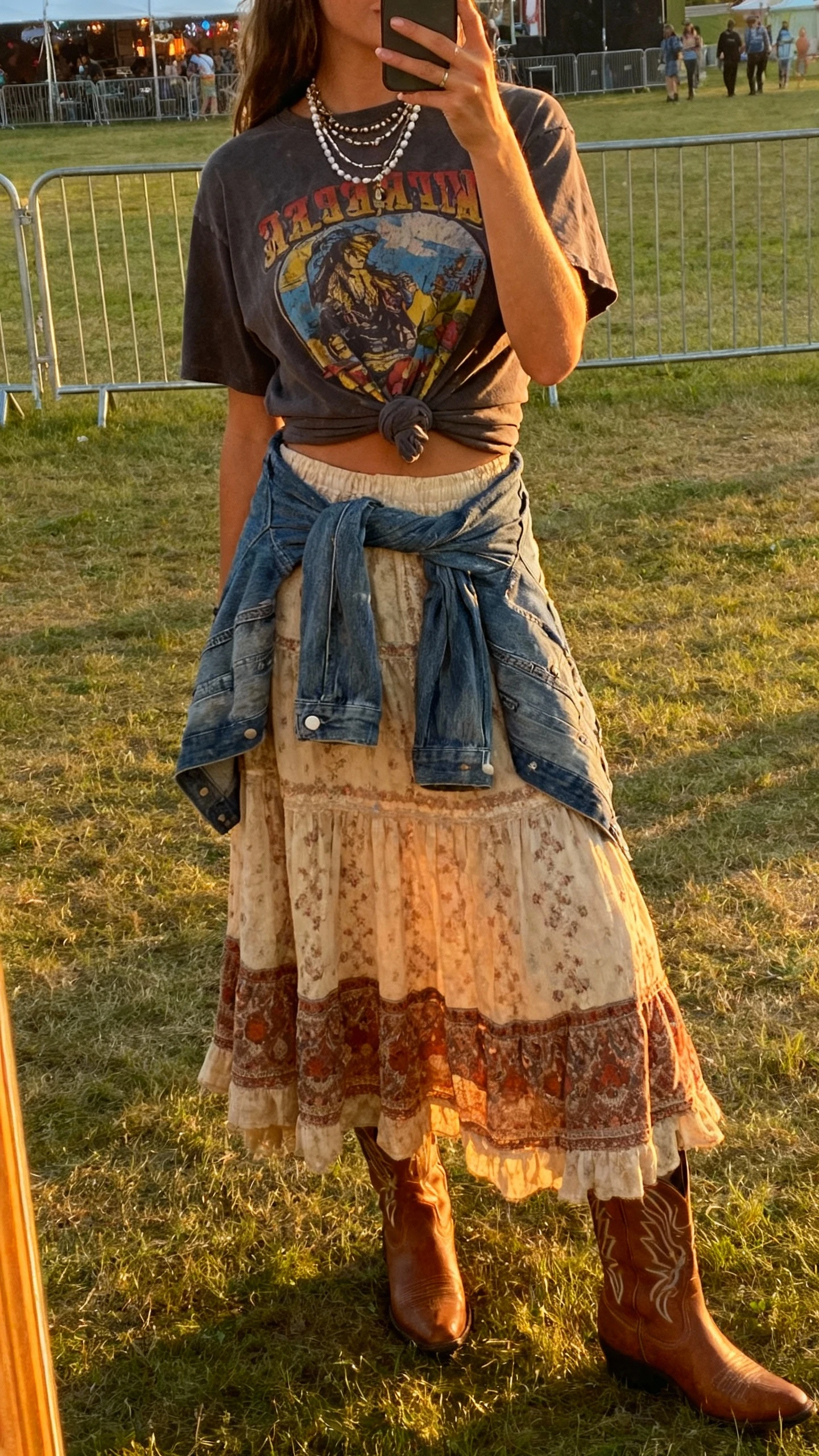 iPhone back-view mirror selfie of a woman wearing a long boho skirt with a vintage graphic tee knotted at the waist, western boots, denim jacket tied around hips, layered necklaces, face not visible, festival lawn setting near a fence, golden-hour daylight, natural iPhone photo quality