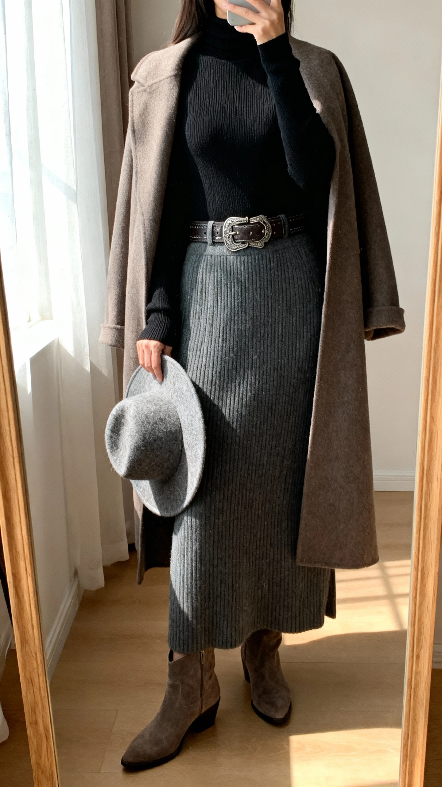 iPhone back-view selfie in a full-length mirror of a woman wearing a charcoal ribbed knit maxi skirt, slim black merino turtleneck, western-inspired belt, suede ankle boots, longline coat draped over shoulders, and a wool fedora in hand; face not visible, soft afternoon window light, natural iPhone photo.