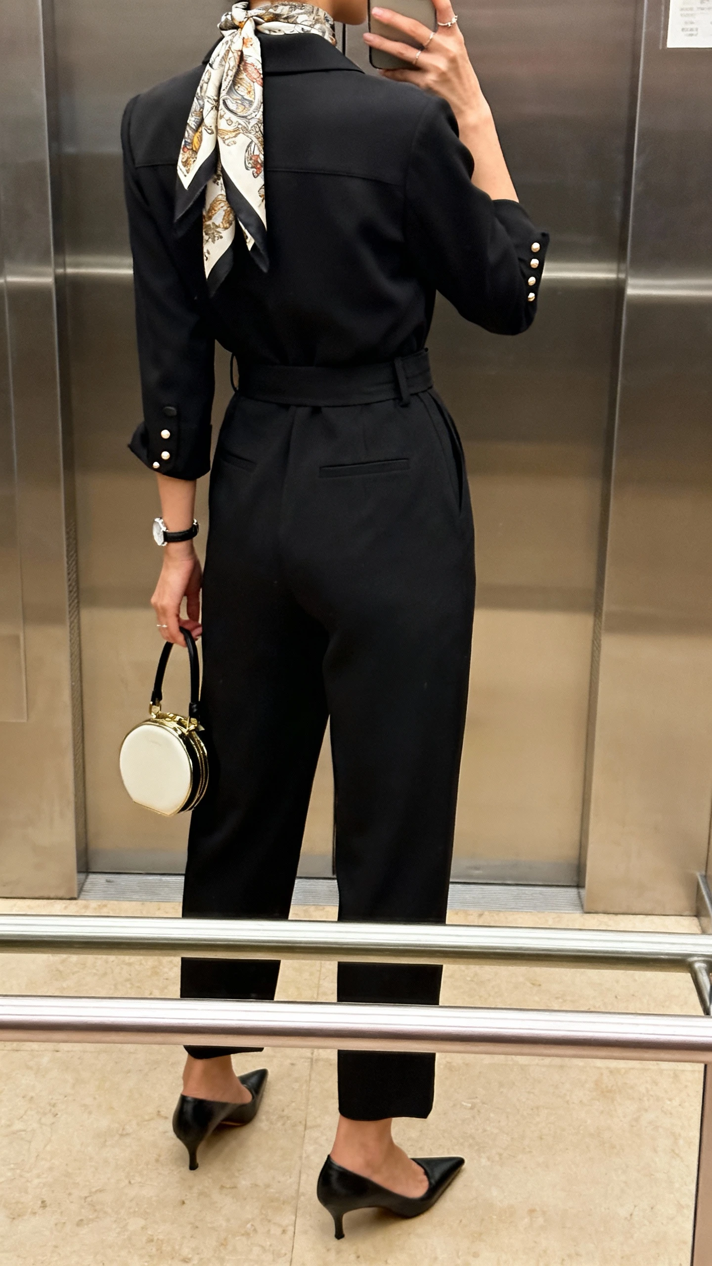 iPhone back-view selfie of a woman in a belted black tailored jumpsuit with a patterned silk scarf tied at the neck, pointed-toe pumps, understated studs, a slim watch, and a compact top-handle bag, face not visible, elevator mirror, warm indoor lighting.