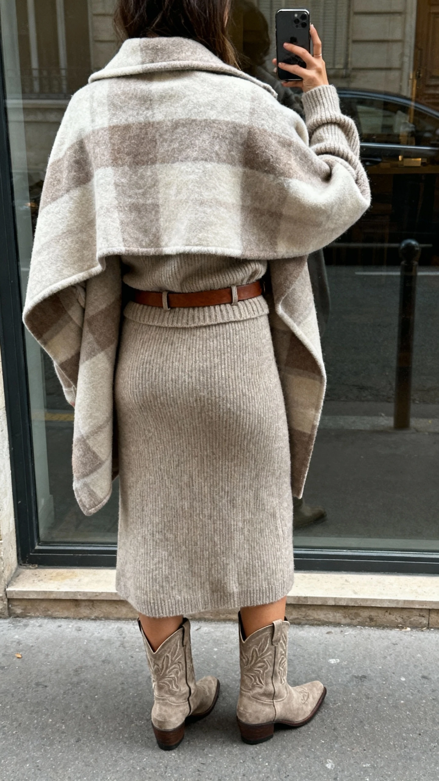 iPhone back-view selfie of a woman in a blanket coat over a tonal knit set (matching sweater and skirt) with western-inspired ankle boots and a simple leather belt, face not visible, street reflection window, overcast daylight, natural iPhone photo quality.