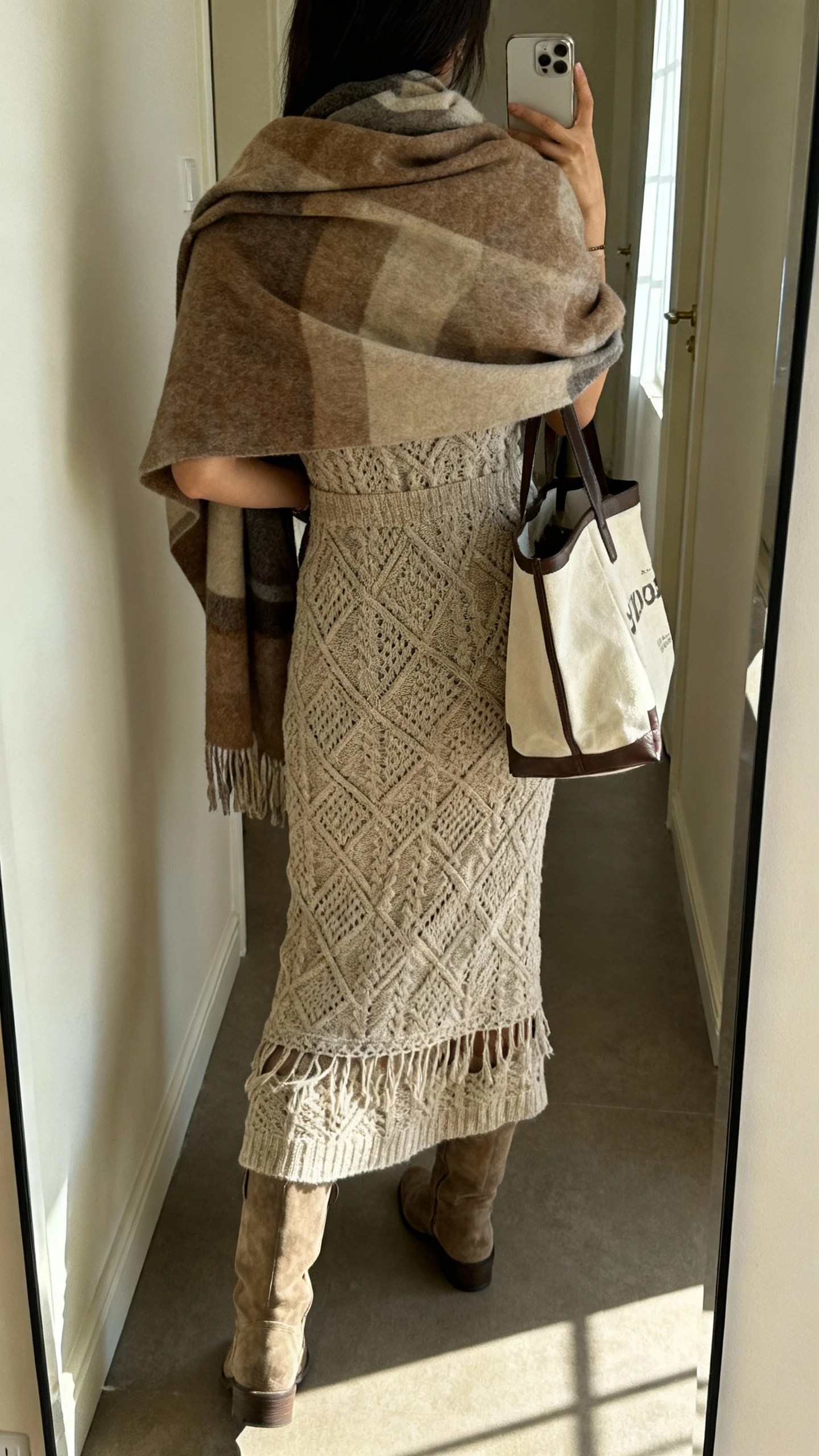 iPhone back-view selfie of a woman in a boho knit dress with tall boots and a large blanket scarf draped over her shoulders, tote bag, face not visible, hallway mirror, soft afternoon window light, natural iPhone photo quality.