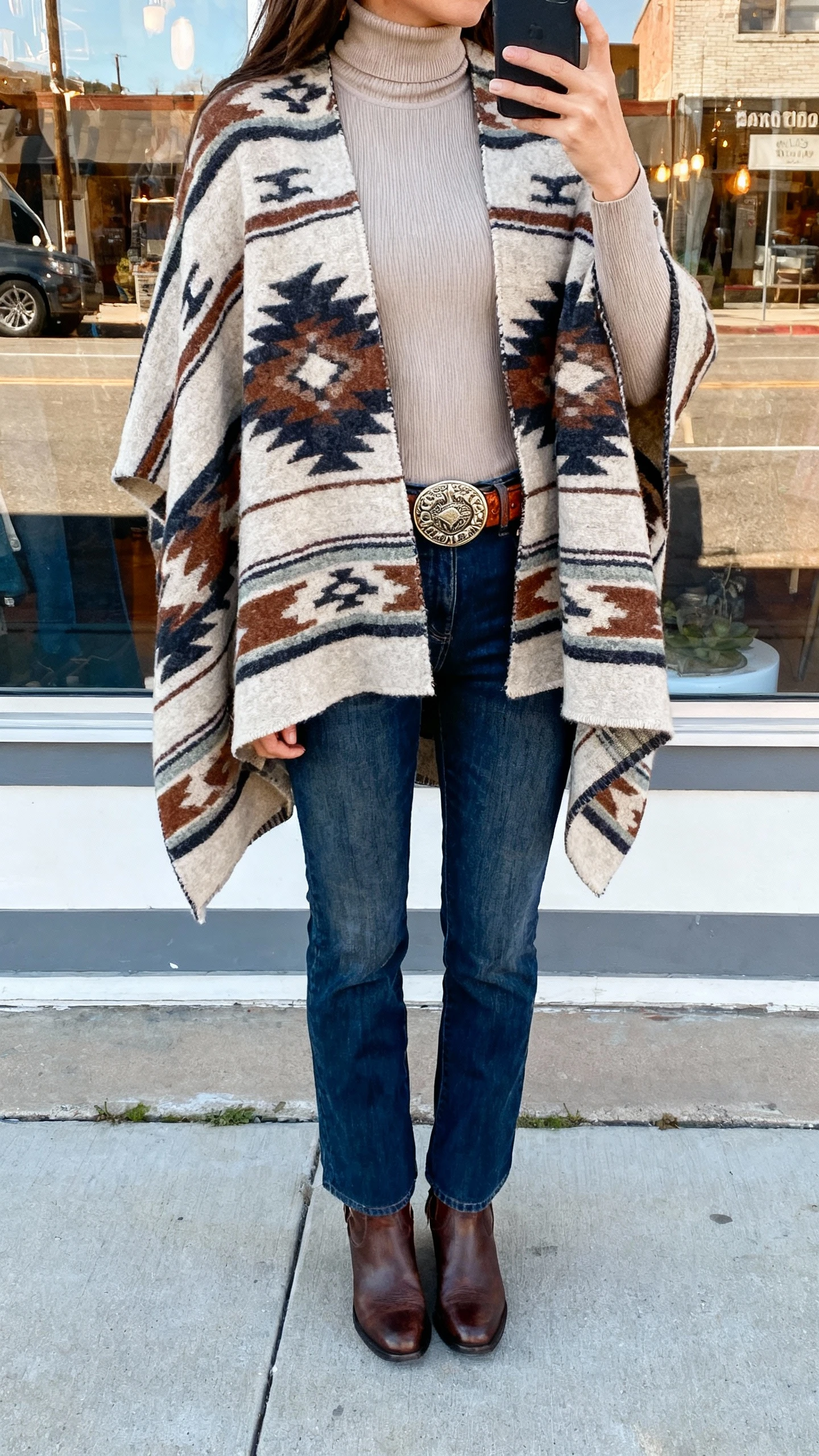 iPhone back-view selfie of a woman in a Southwestern-pattern wool poncho over a fitted neutral turtleneck, straight-leg jeans, leather ankle boots, tooled leather belt with bold buckle, face not visible, street reflection in a shop window, natural daylight, casual iPhone photo.