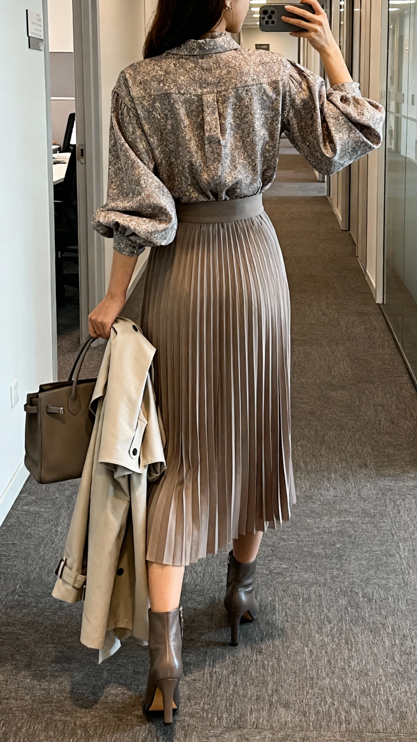 iPhone back-view selfie of a woman in a statement blouse with volume sleeves tucked into a midi pleated skirt, heeled ankle boots, lightweight trench over arm, structured handbag, face not visible, office hallway with indoor lighting, natural iPhone quality.