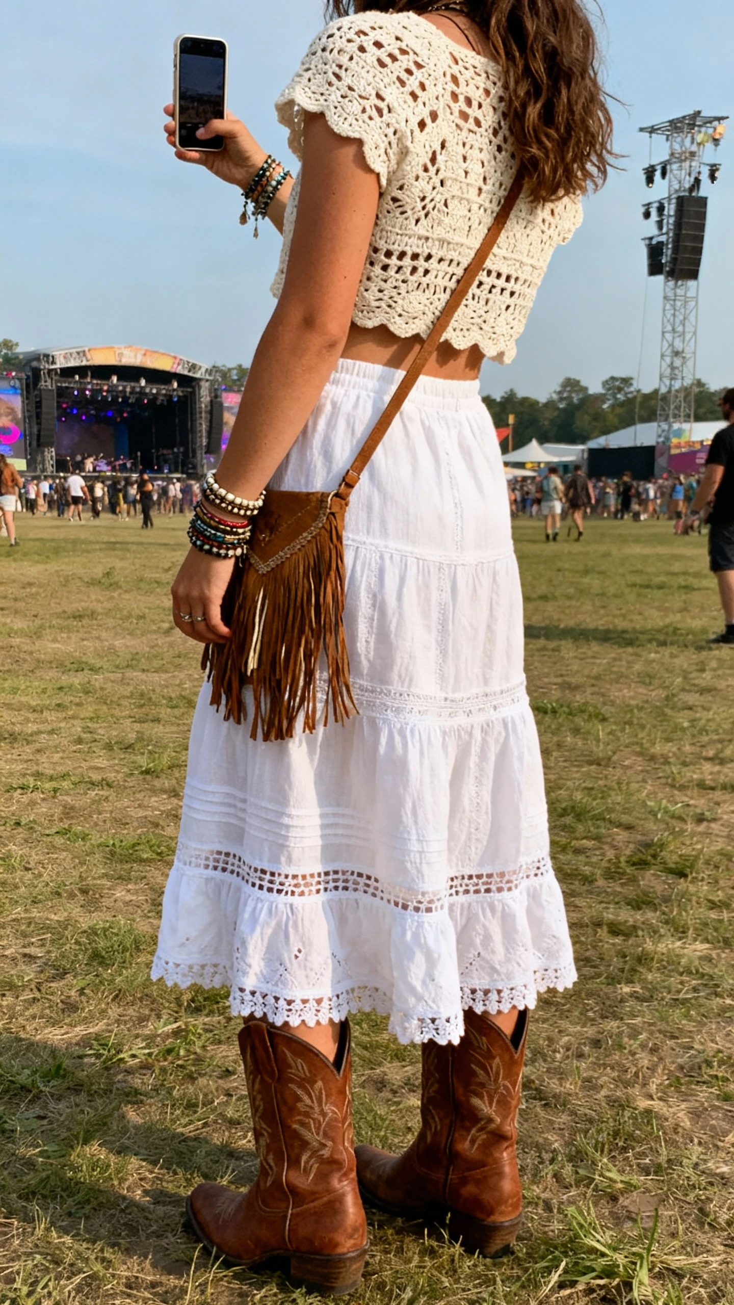 iPhone back-view selfie of a woman in a white boho skirt paired with a cream crochet top, brown cowboy boots, layered bracelets, fringe crossbody, face not visible, taken at a music festival field with daylight, casual iPhone photo.