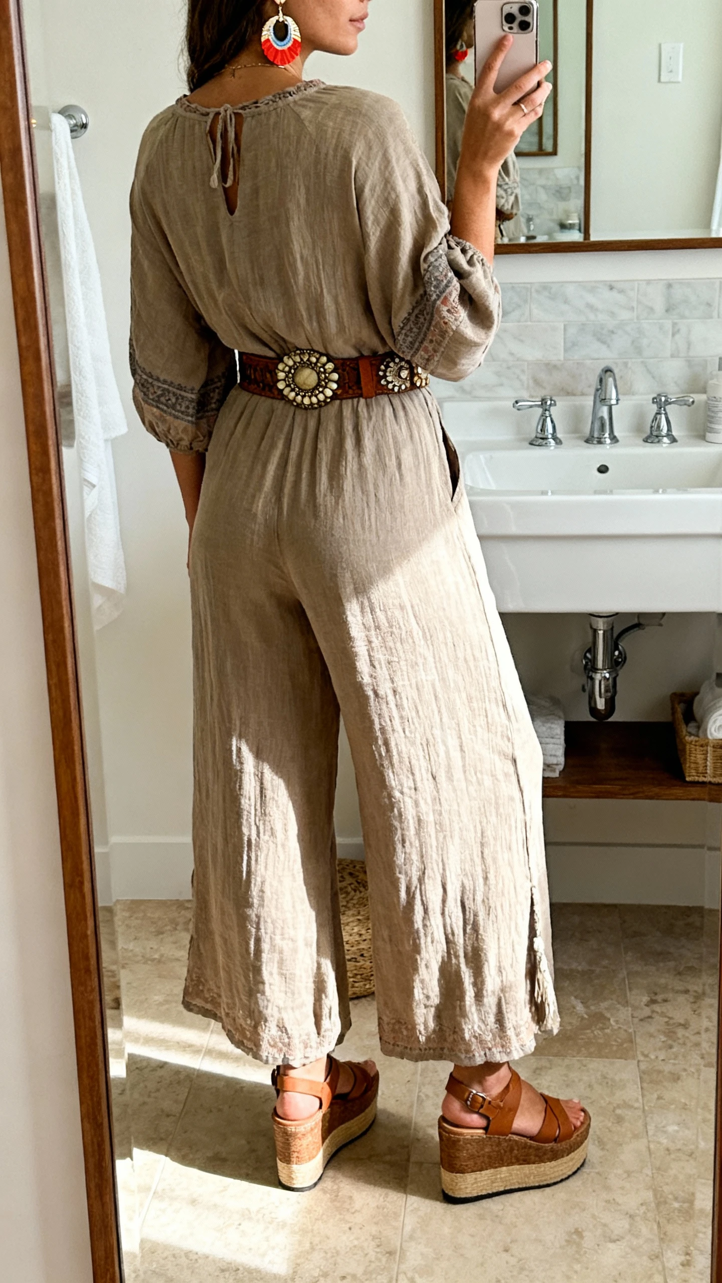 iPhone back-view selfie of a woman in a wide-leg boho jumpsuit cinched with a statement belt, platform sandals, bold earrings, face not visible, taken in a bathroom mirror under natural morning light, casual iPhone quality.