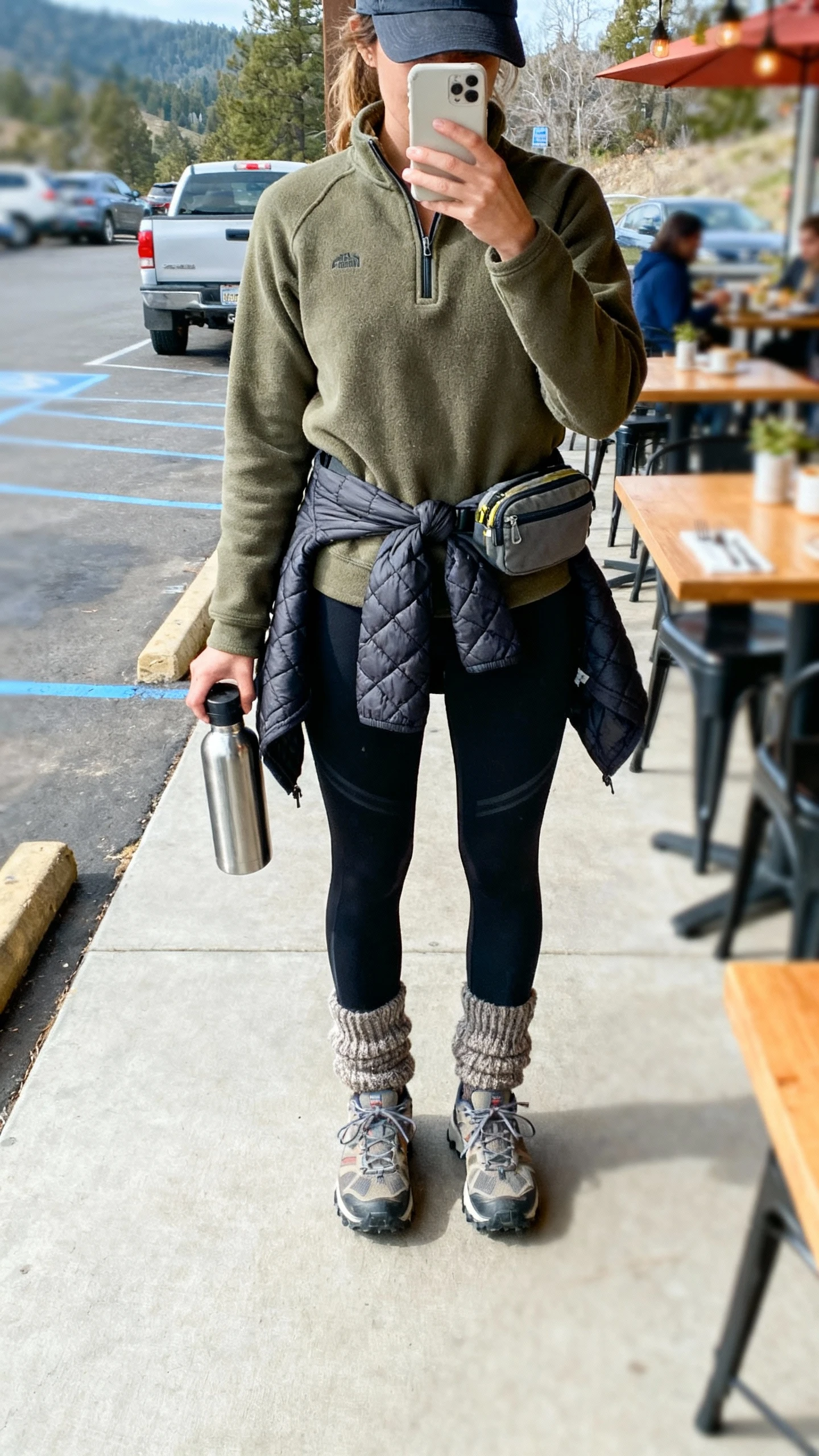 iPhone back-view selfie of a woman in hike-to-brunch athleisure: olive half-zip fleece over black performance leggings, trail sneakers, quilted vest tied at waist, chunky socks, baseball cap, small belt bag, stainless water bottle in hand, face not visible, trailhead parking lot to cafe patio, bright daylight, casual iPhone photo.