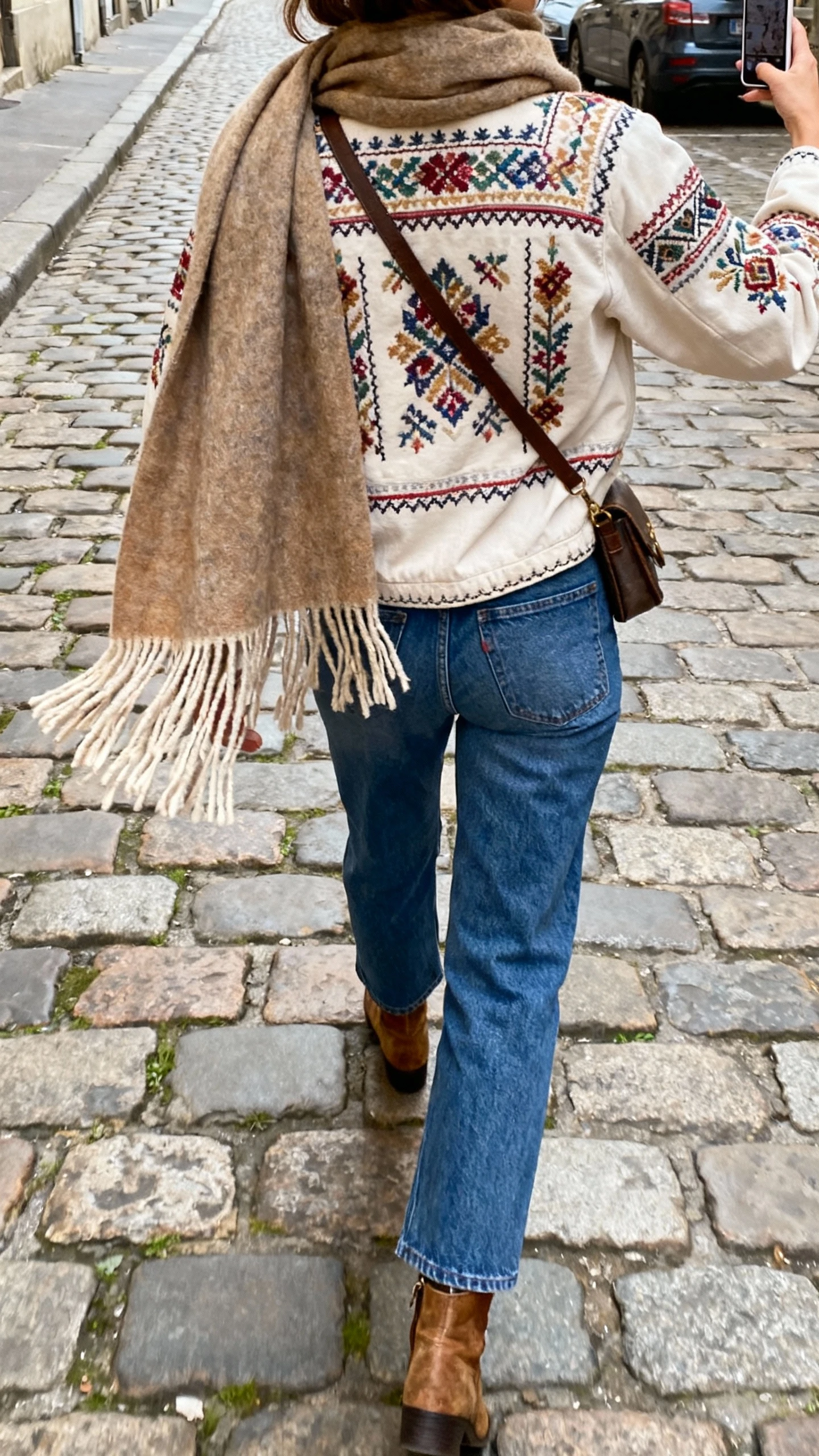 iPhone back-view selfie of a woman wearing a folk embroidered jacket with straight blue denim, a big fringed scarf, ankle boots, crossbody bag, face not visible, walking on a cobblestone street, natural daylight, casual iPhone photo