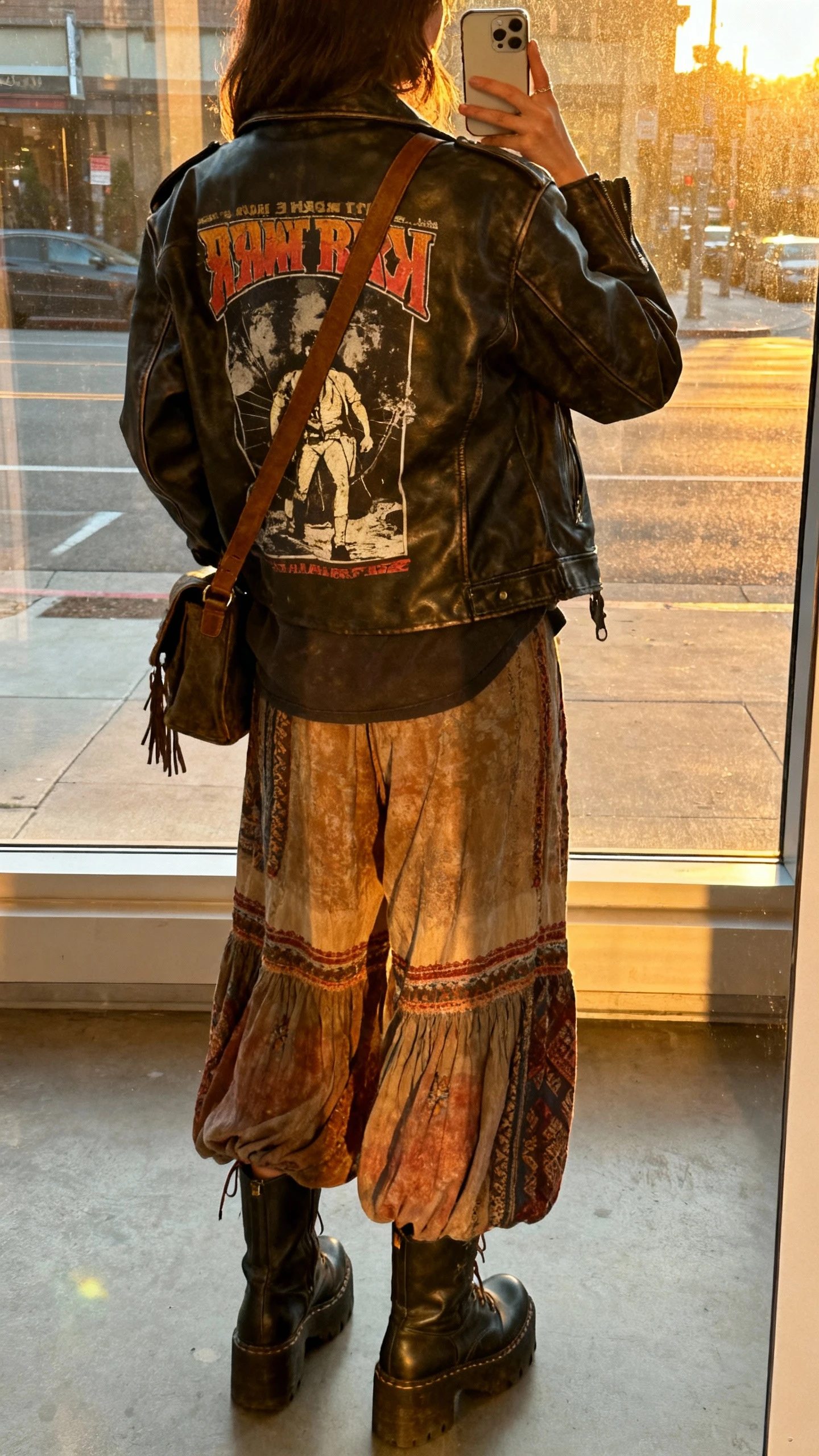 iPhone back-view selfie of a woman wearing a leather jacket over a vintage band tee with flowy boho pants and chunky boots, crossbody bag, face not visible, city street window reflection at golden hour, natural iPhone photo quality.