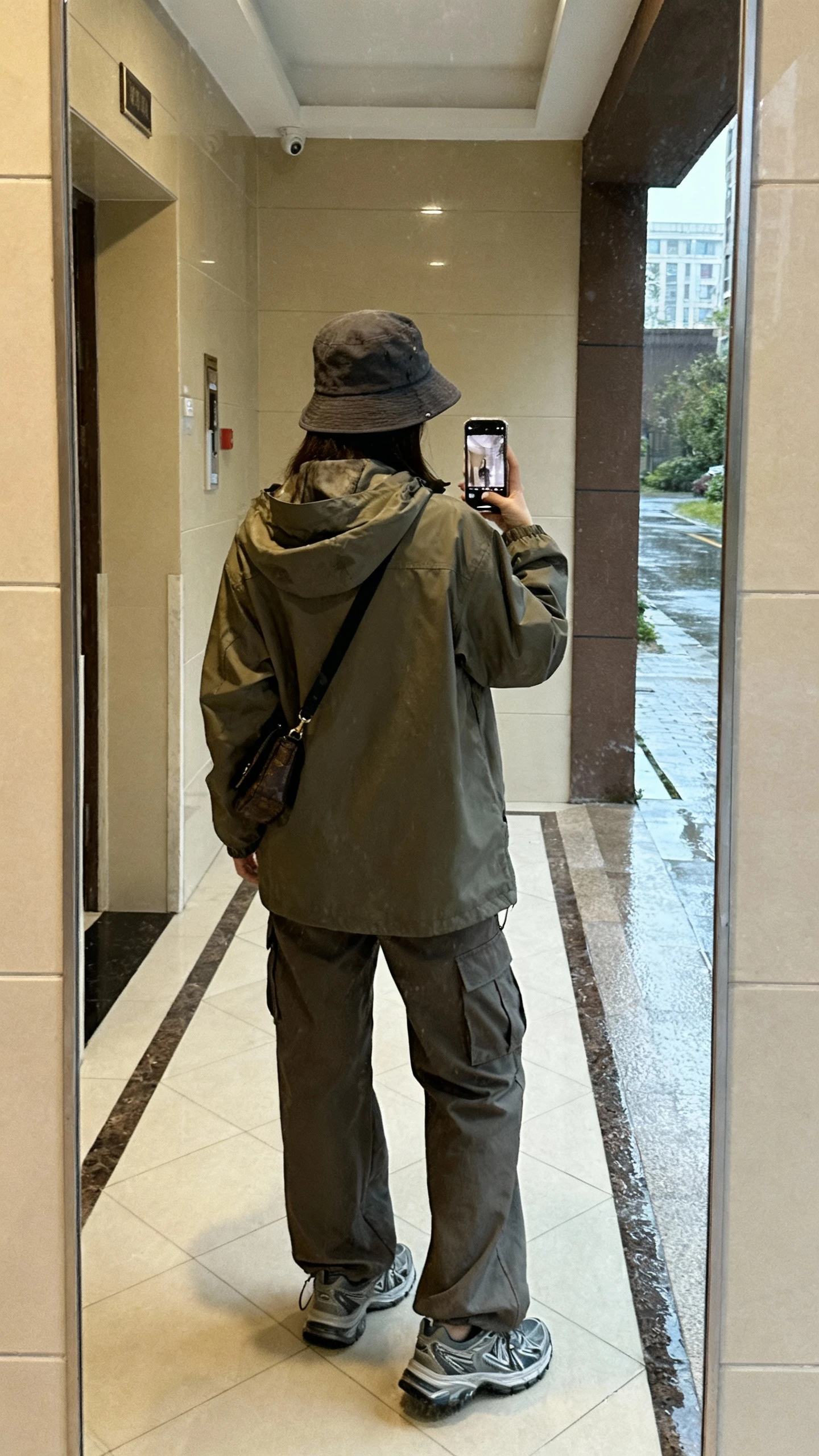 iPhone back-view selfie of a woman wearing a rain-ready parka with hood up, relaxed cargo trousers, waterproof sneakers, bucket hat, crossbody; face not visible, shot in apartment lobby mirror with wet pavement visible, soft indoor lighting, casual iPhone look.