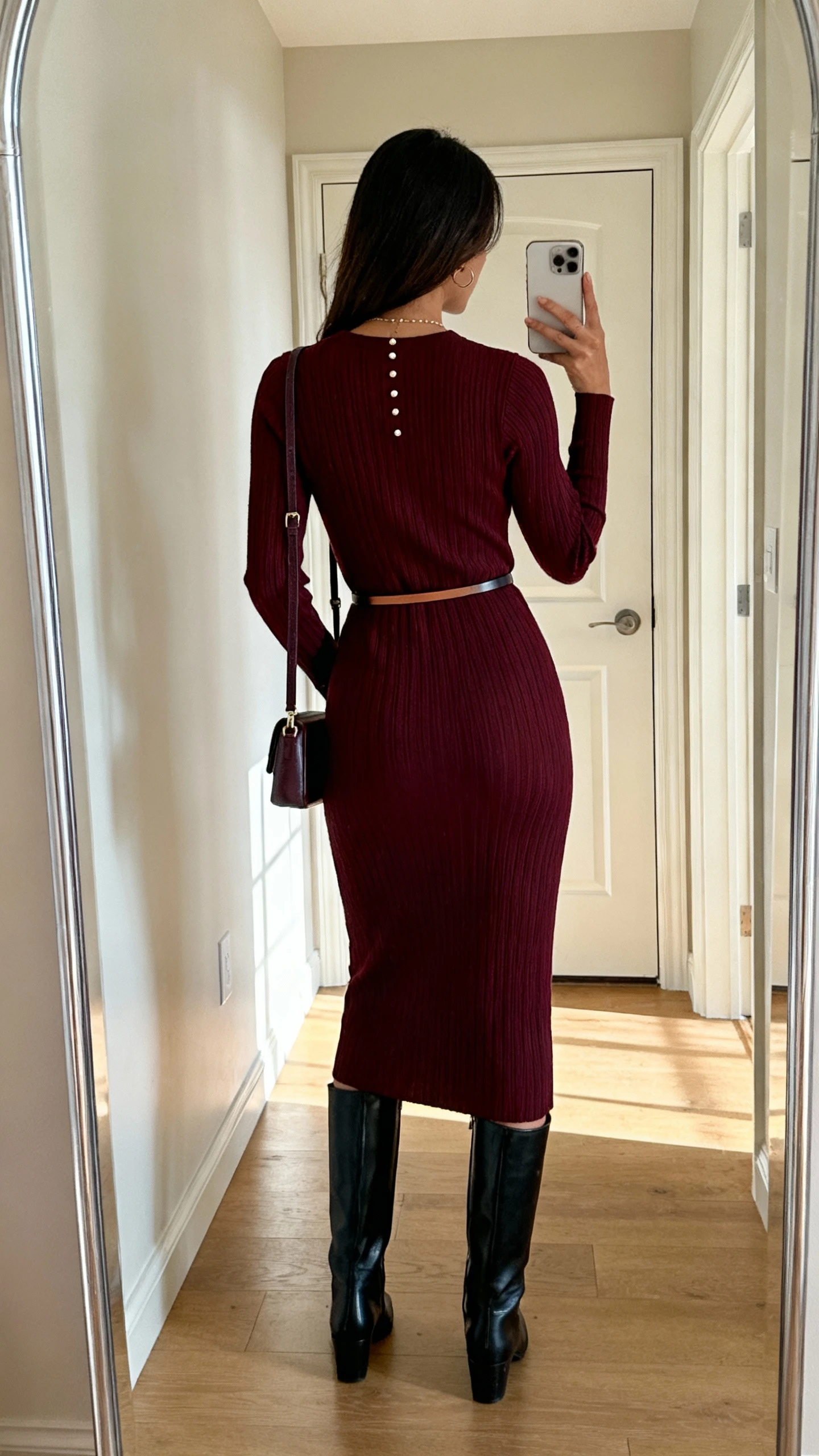 iPhone back-view selfie of a woman wearing a ribbed burgundy knit midi dress, black knee-high boots, thin belt, delicate studs, and a small shoulder bag, face not visible, taken in a hallway mirror by the front door, soft afternoon light, casual iPhone photo.