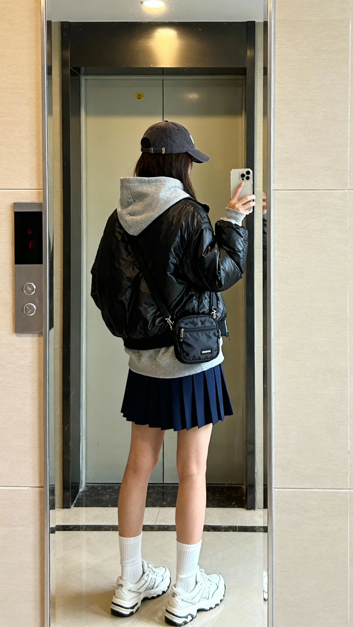 iPhone back-view selfie of a woman wearing a sporty-chic combo: black nylon bomber layered over a grey hoodie, navy pleated skort, white crew socks, retro white sneakers, baseball cap, and a mini nylon crossbody, face not visible, elevator mirror in an apartment building, indoor lighting, casual iPhone quality.