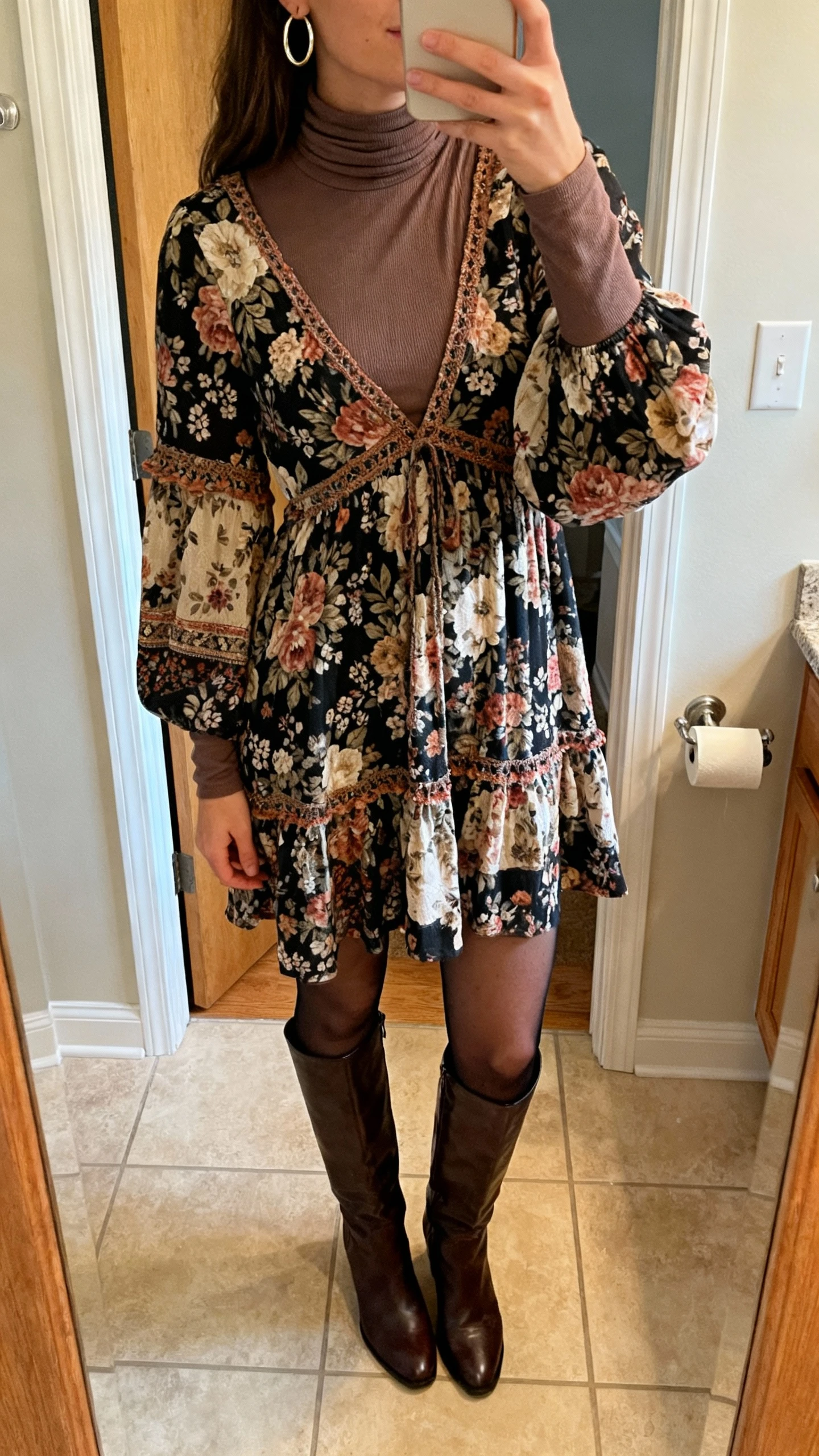 iPhone bathroom mirror selfie of a woman wearing a floral boho dress over a fitted turtleneck, opaque tights, knee-high boots, and hoop earrings, face not visible, cropped at shoulders, warm indoor lighting, natural iPhone aesthetic.
