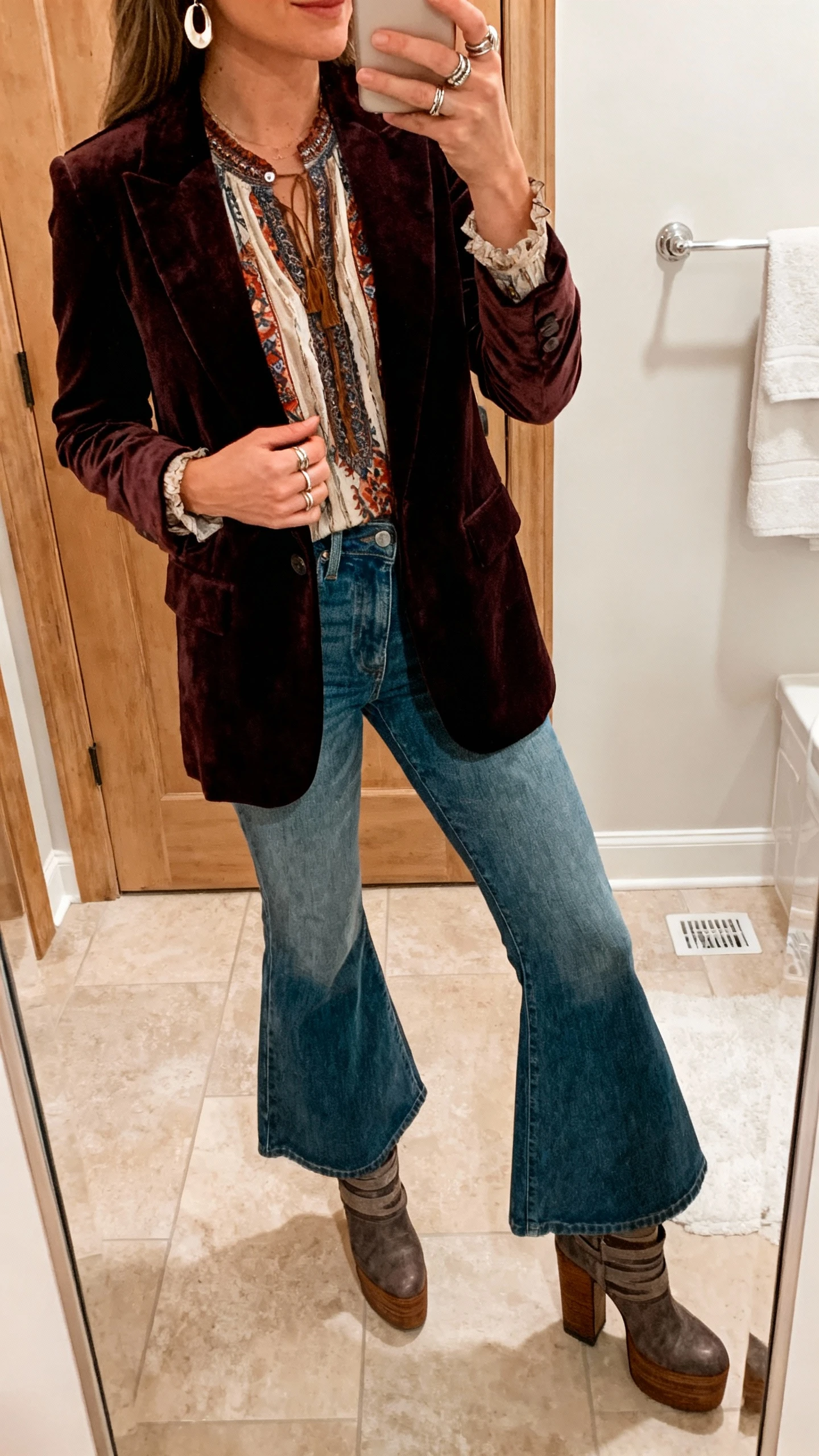 iPhone bathroom mirror selfie of a woman wearing a velvet blazer over a boho blouse with flare jeans and stacked-heel boots, layered rings and earrings, face not visible, warm indoor lighting, natural iPhone aesthetic