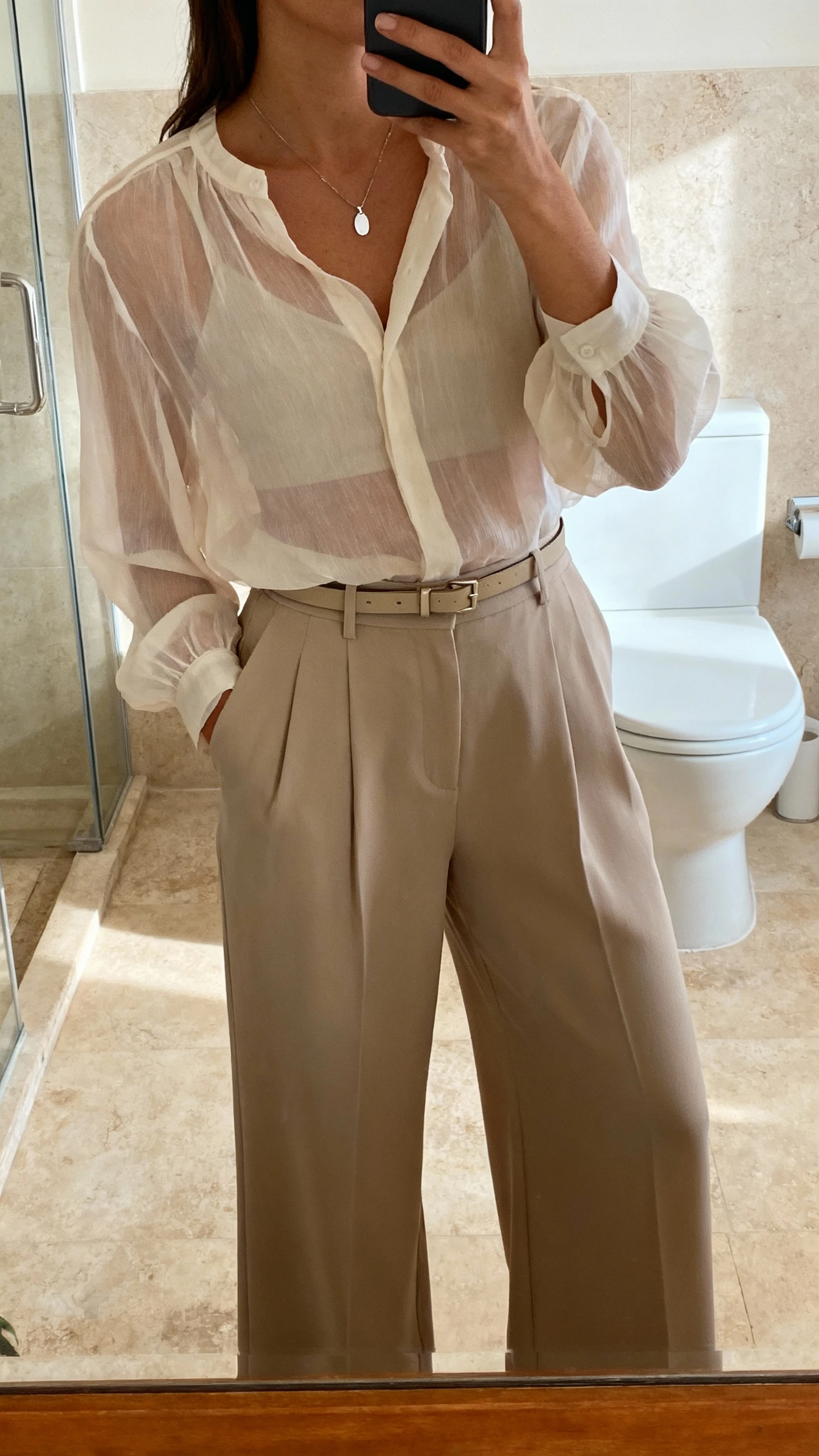 iPhone bathroom mirror selfie of a woman wearing wide-leg trousers with a sheer blouse, delicate pendant, and slim belt, face not visible, cropped at shoulders, warm indoor lighting, natural iPhone photo quality.