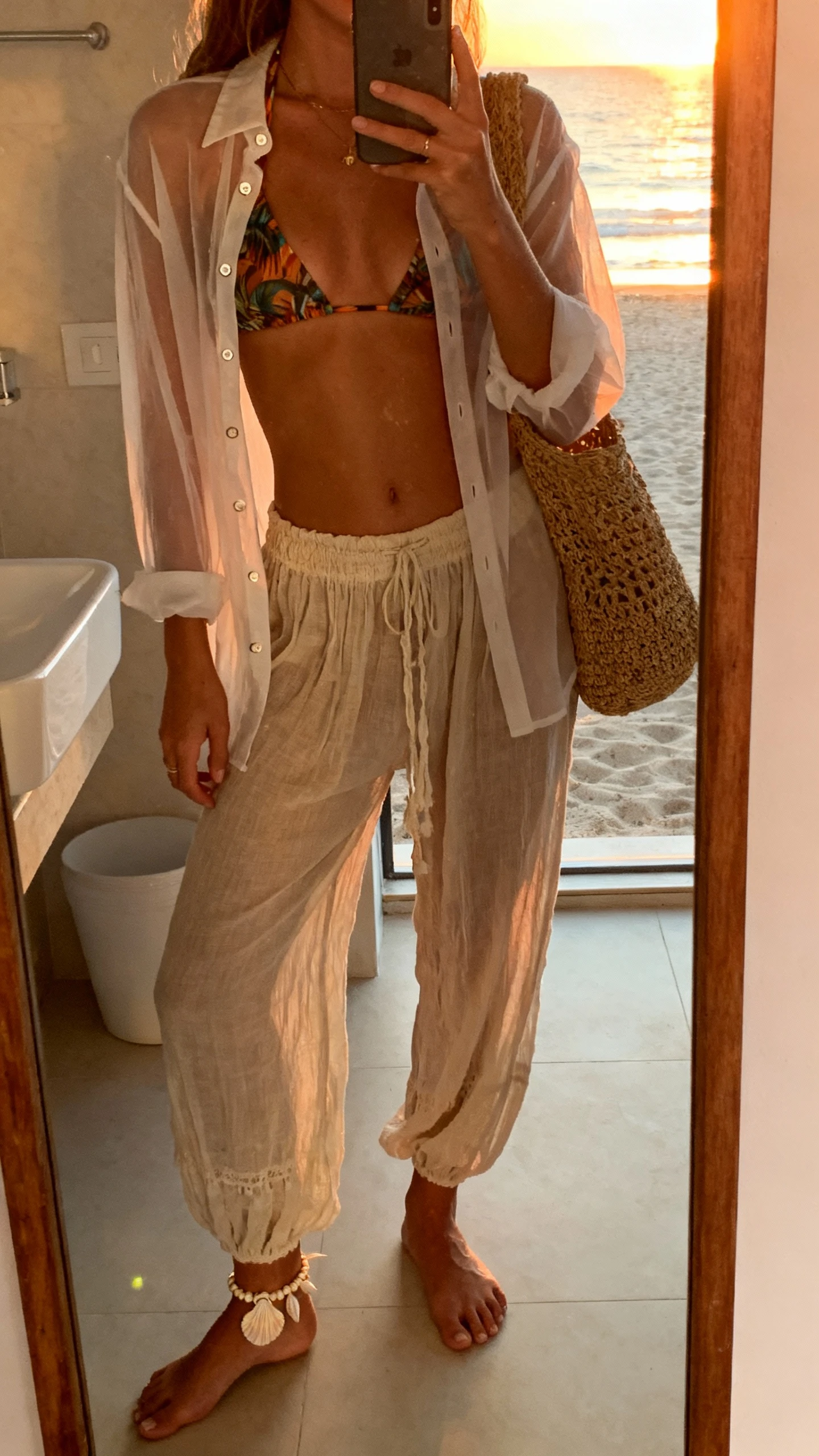 iPhone beach bathroom mirror selfie of a woman wearing a bikini top layered under a sheer button-up shirt with airy boho pants, shell anklet, woven tote, face not visible, sunset glow through window, natural iPhone photo quality.