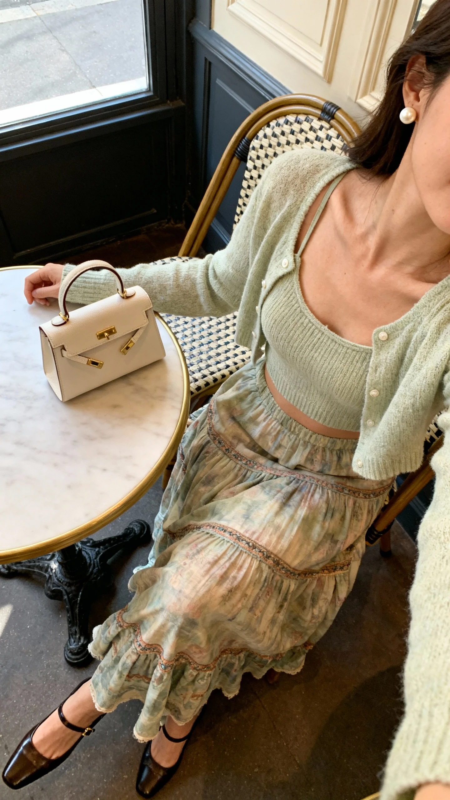 iPhone café table selfie angled down showing a woman wearing a long boho skirt with a matching cardigan set (camisole + cropped cardigan), slingback heels, pearl studs, small top-handle bag, face not visible, Parisian-style café interior, morning window light, natural iPhone photo quality