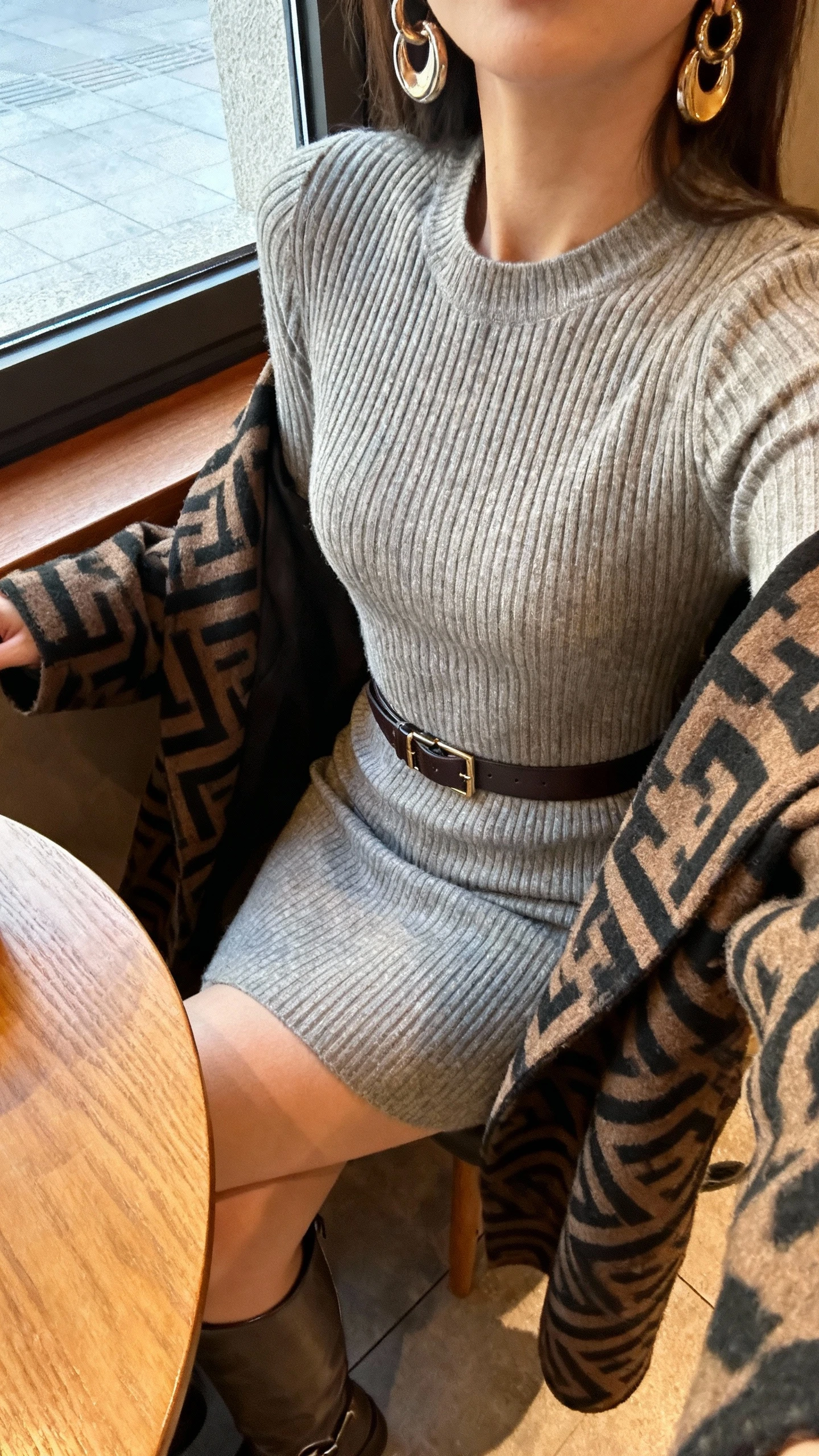 iPhone cafe table selfie cropped at neck showing a woman wearing a ribbed sweater dress cinched with a wide belt, statement patterned coat draped over shoulders, knee-high boots, and bold earrings, face not visible, warm cafe lighting by window, natural iPhone photo quality