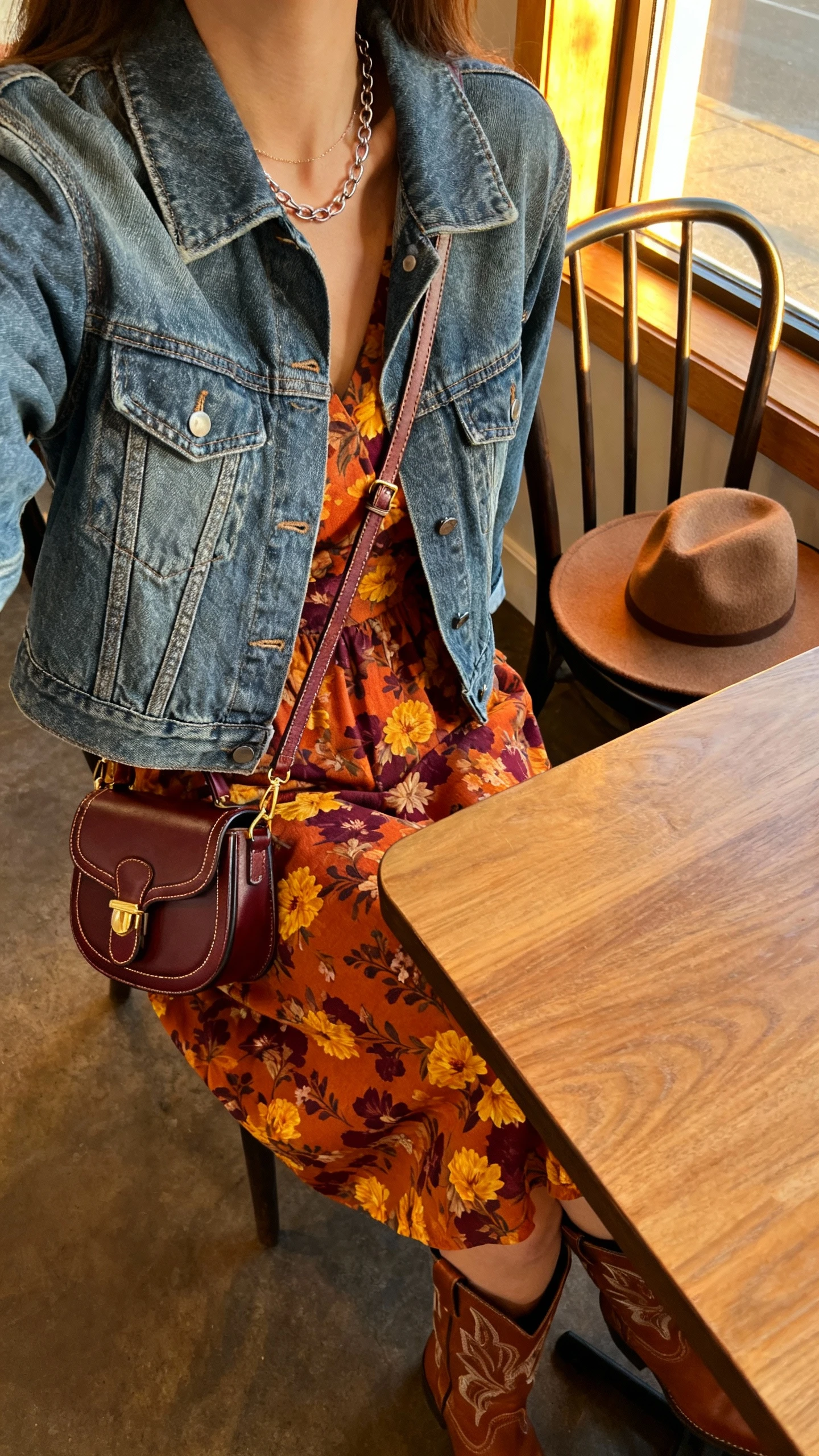 iPhone cafe table selfie cropped at shoulders of a woman wearing a mid-wash denim jacket (classic or slightly cropped) over a fall floral midi dress (burnt orange/marigold/plum), ankle or western-inspired boots, delicate chain necklace, crossbody saddle or mini top-handle bag, optional wide-brim felt hat on chair, face not visible, golden hour window light, natural iPhone photo.