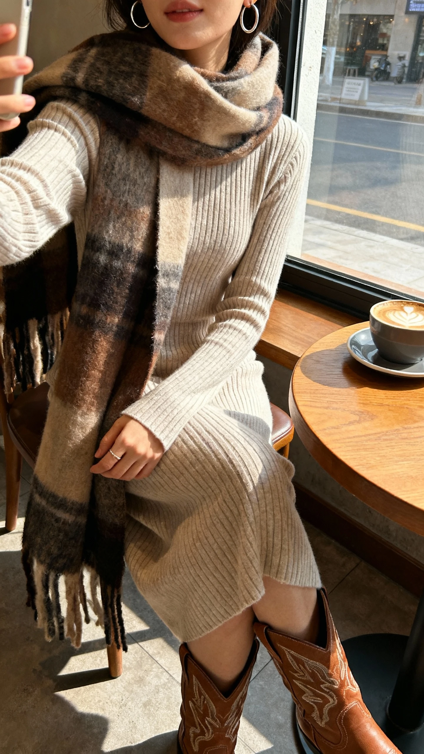 iPhone café table selfie cropped at shoulders showing a woman in a ribbed sweater dress with a large blanket scarf, Western boots, and hoop earrings, face not visible, sitting by a window in a coffee shop, morning light, casual iPhone photo.