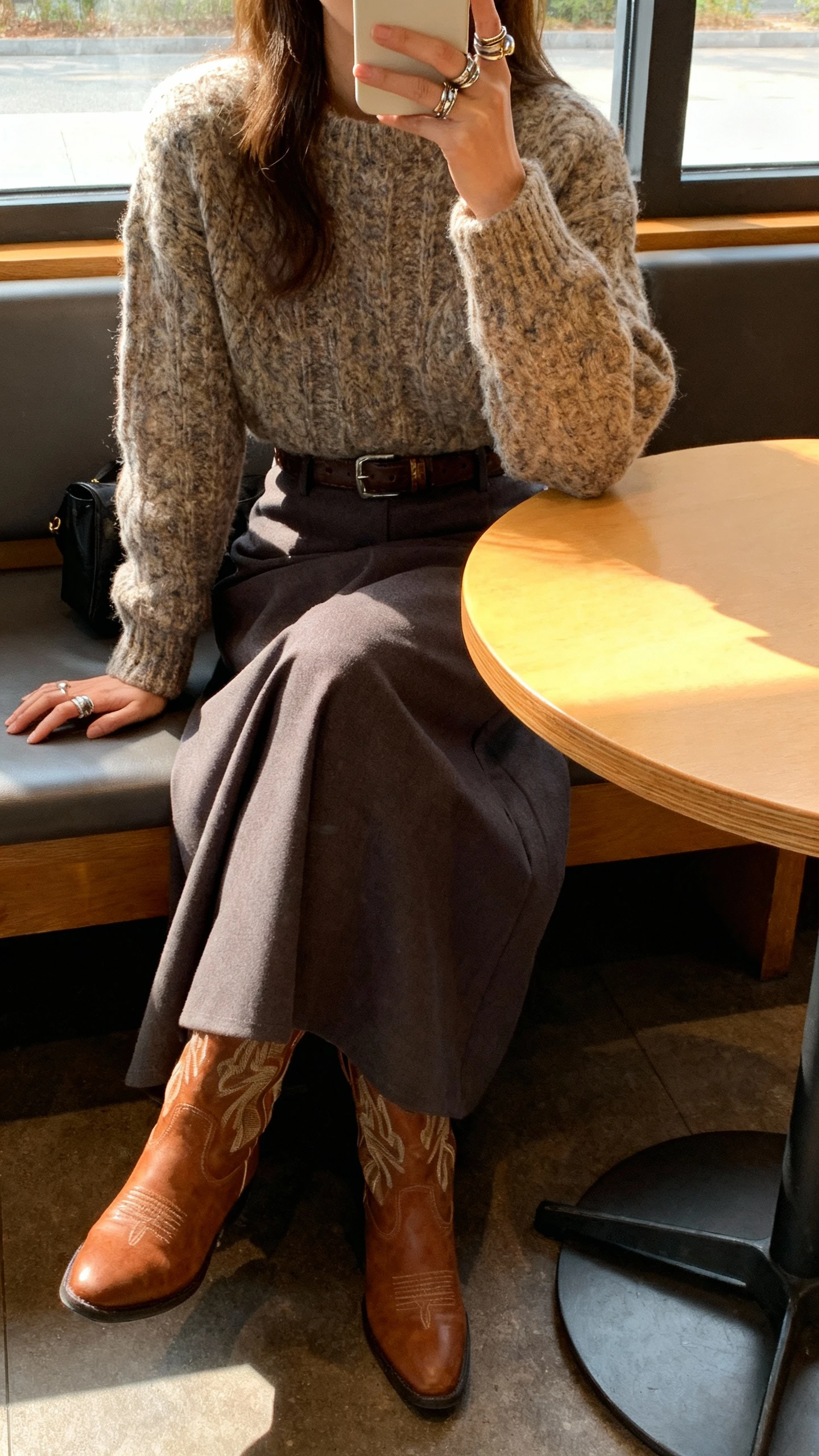 iPhone café table selfie cropped at shoulders showing a woman wearing a chunky sweater over a maxi skirt with Western boots, stacked rings, and a belt, face not visible, window light in coffee shop, natural iPhone photo quality