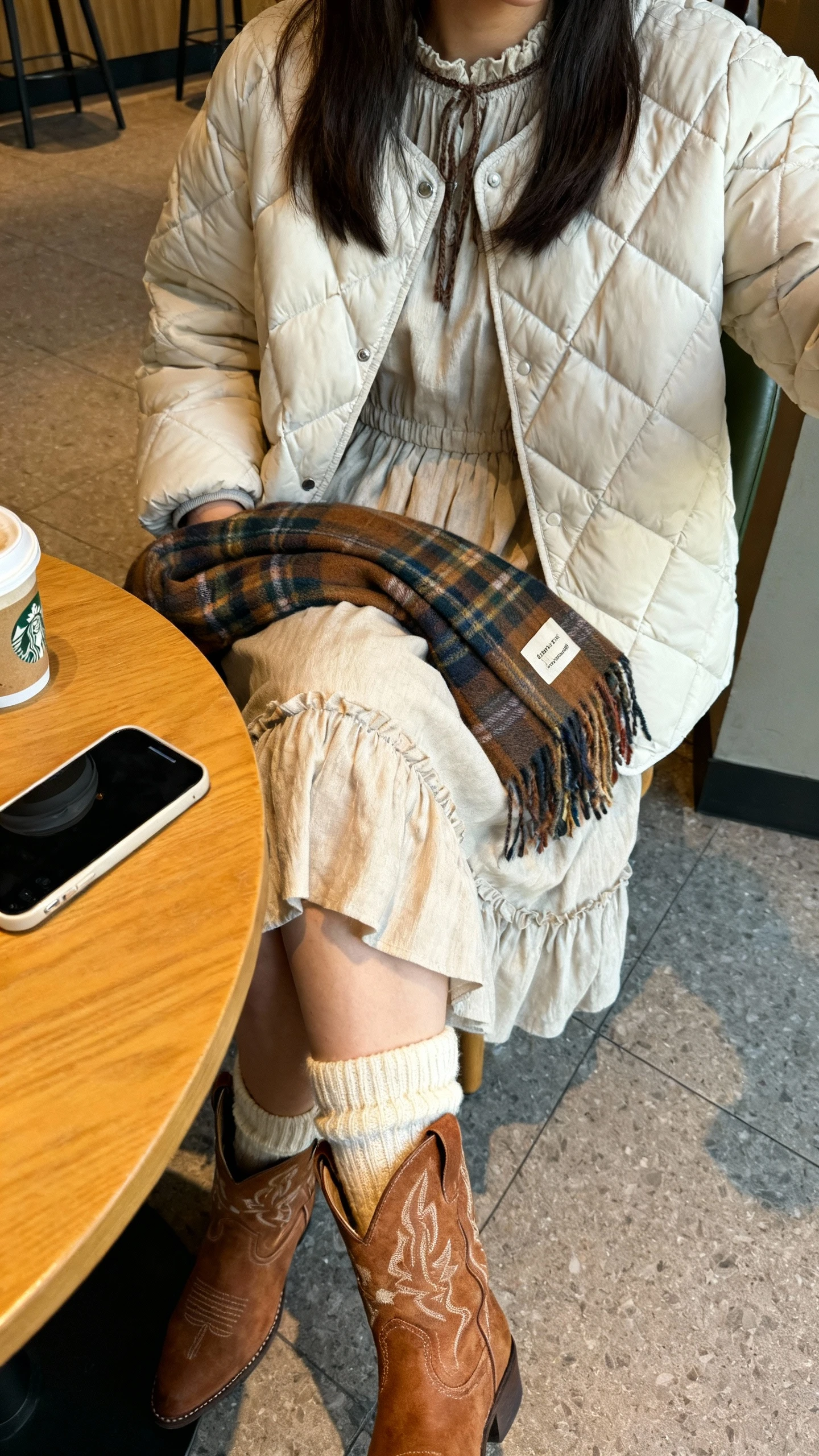 iPhone cafe tabletop selfie (downward angle) showing a woman seated in a quilted puffer jacket over a prairie dress, wool socks peeking from western booties, and a plaid scarf on lap, face not visible, cozy coffee shop with natural daylight, natural iPhone photo quality.