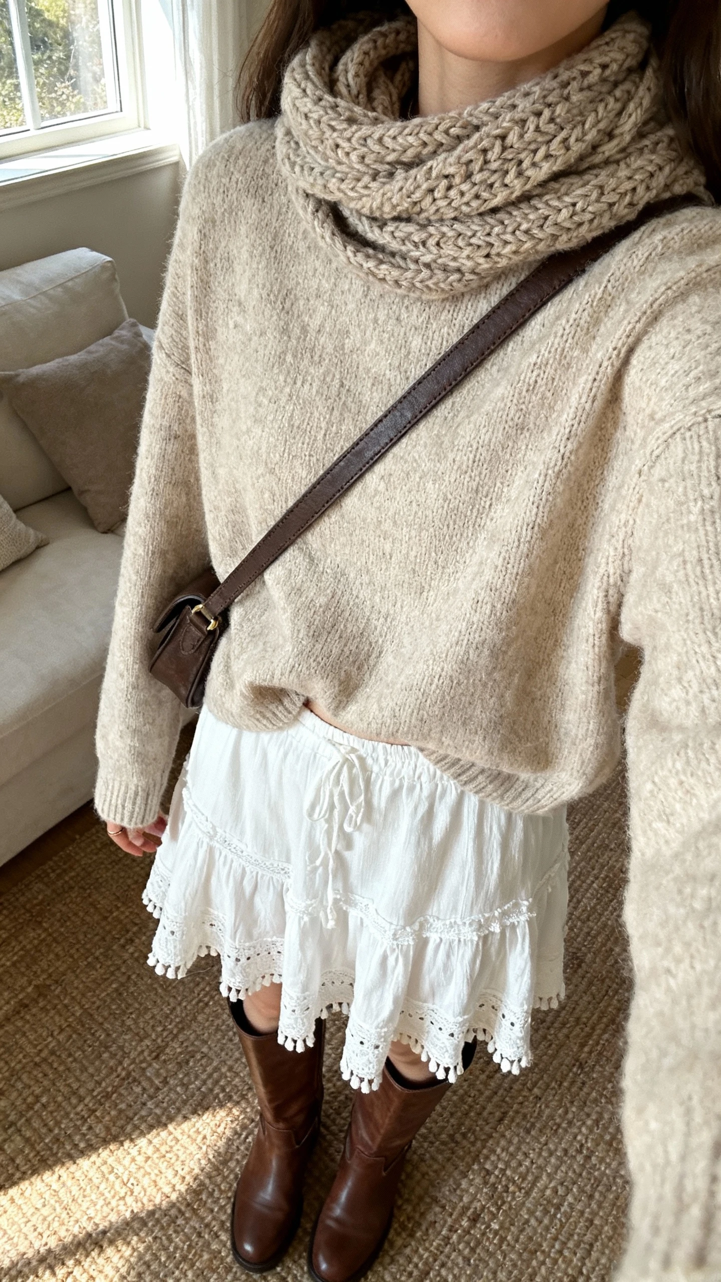 iPhone couch selfie cropped at neck showing a woman in a white boho skirt, cozy oatmeal knit sweater half-tucked, tall brown boots, knit scarf, crossbody bag, face not visible, living room with window light, natural iPhone aesthetic.