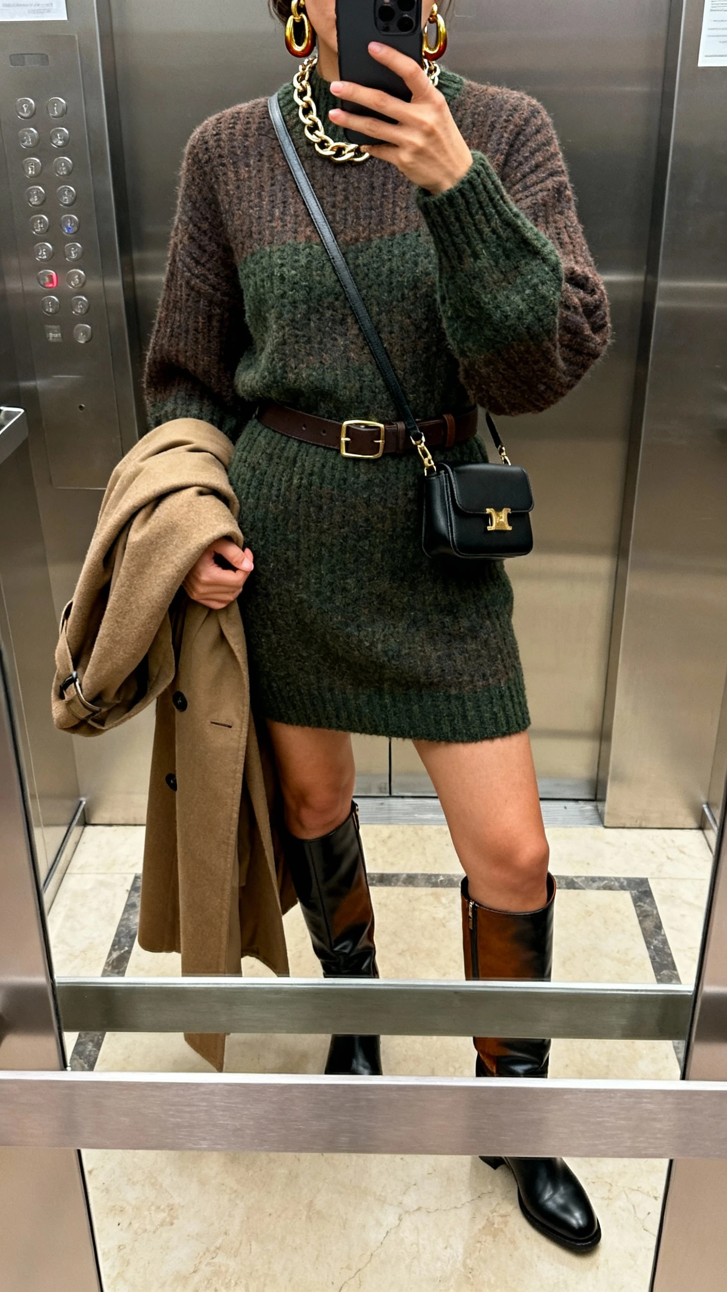 iPhone elevator mirror selfie of a woman in a knit sweater dress (charcoal/chocolate/deep green), knee-high or over-the-knee leather boots (black/brown), optional waist belt, trench coat or long wool coat draped over arm, crossbody or mini shoulder bag, bold earrings or chunky chain, face not visible, indoor lighting, natural iPhone quality.
