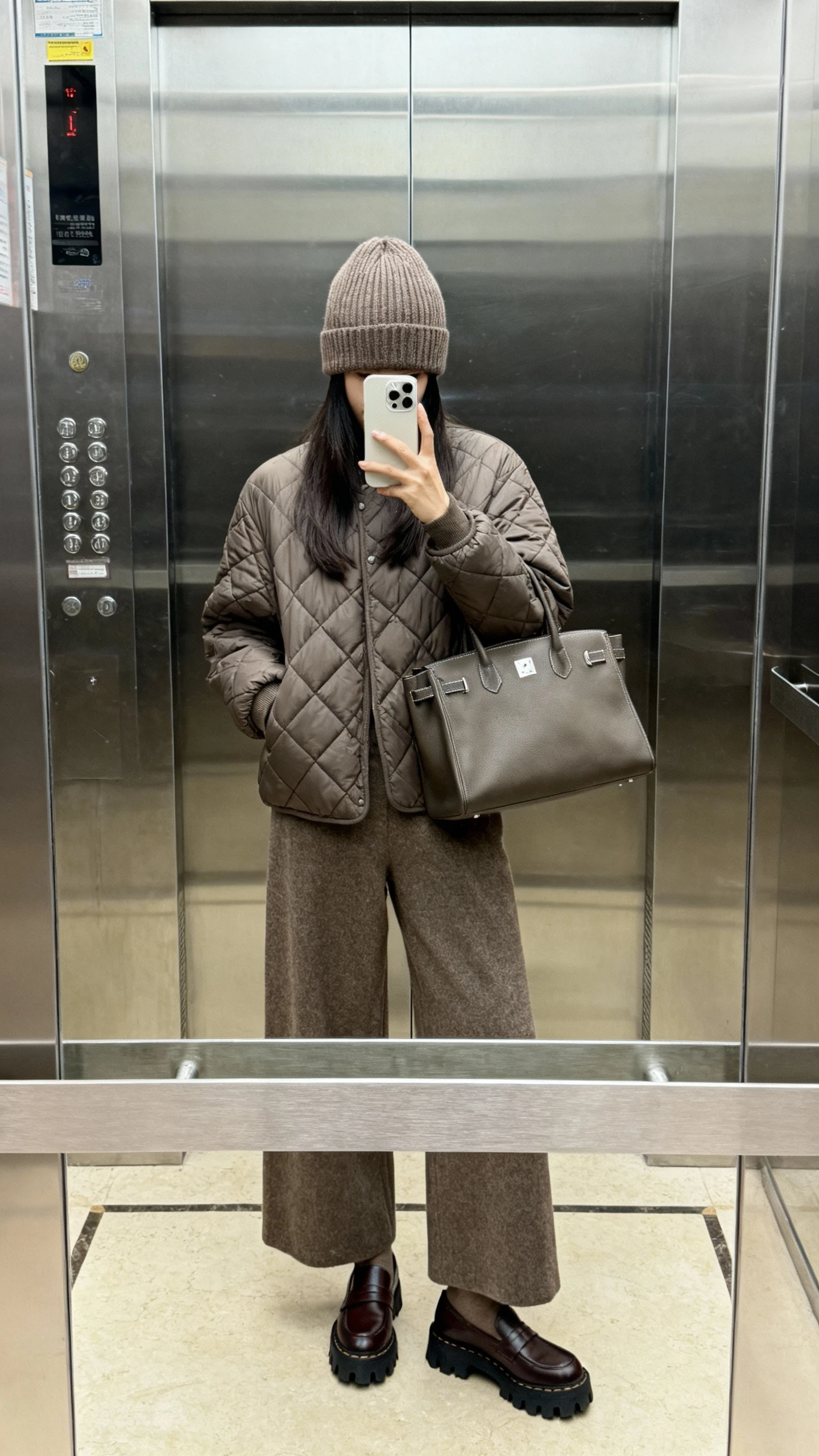iPhone elevator mirror selfie of a woman in a quilted jacket, wide-leg wool trousers, ribbed beanie, lug-sole loafers, and a structured tote, face not visible, indoor lighting with soft reflections, natural iPhone photo.