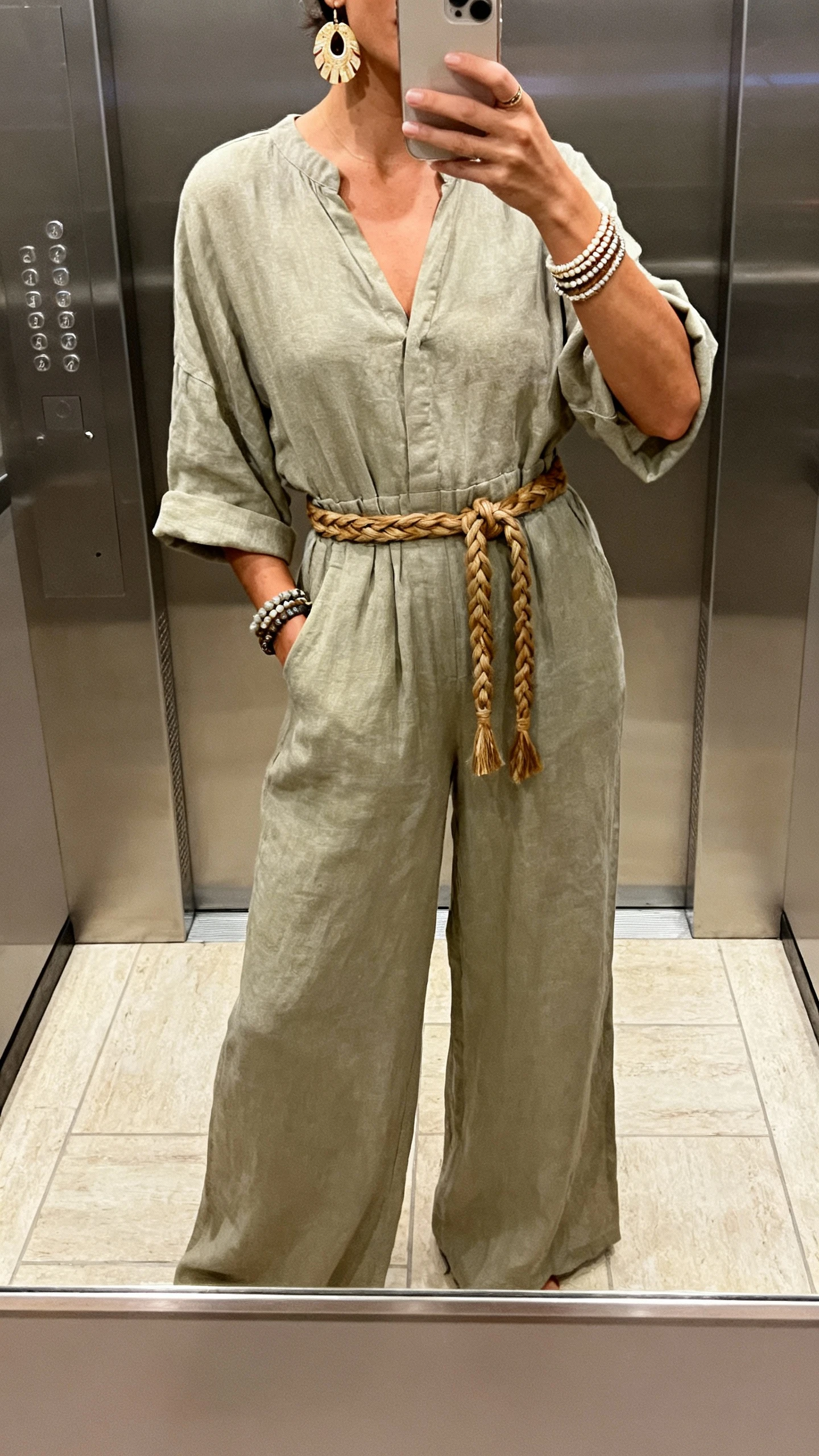 iPhone elevator mirror selfie of a woman in a relaxed jumpsuit with a braided belt, statement earrings, and stacked bracelets, face not visible, indoor lighting, natural iPhone photo quality.