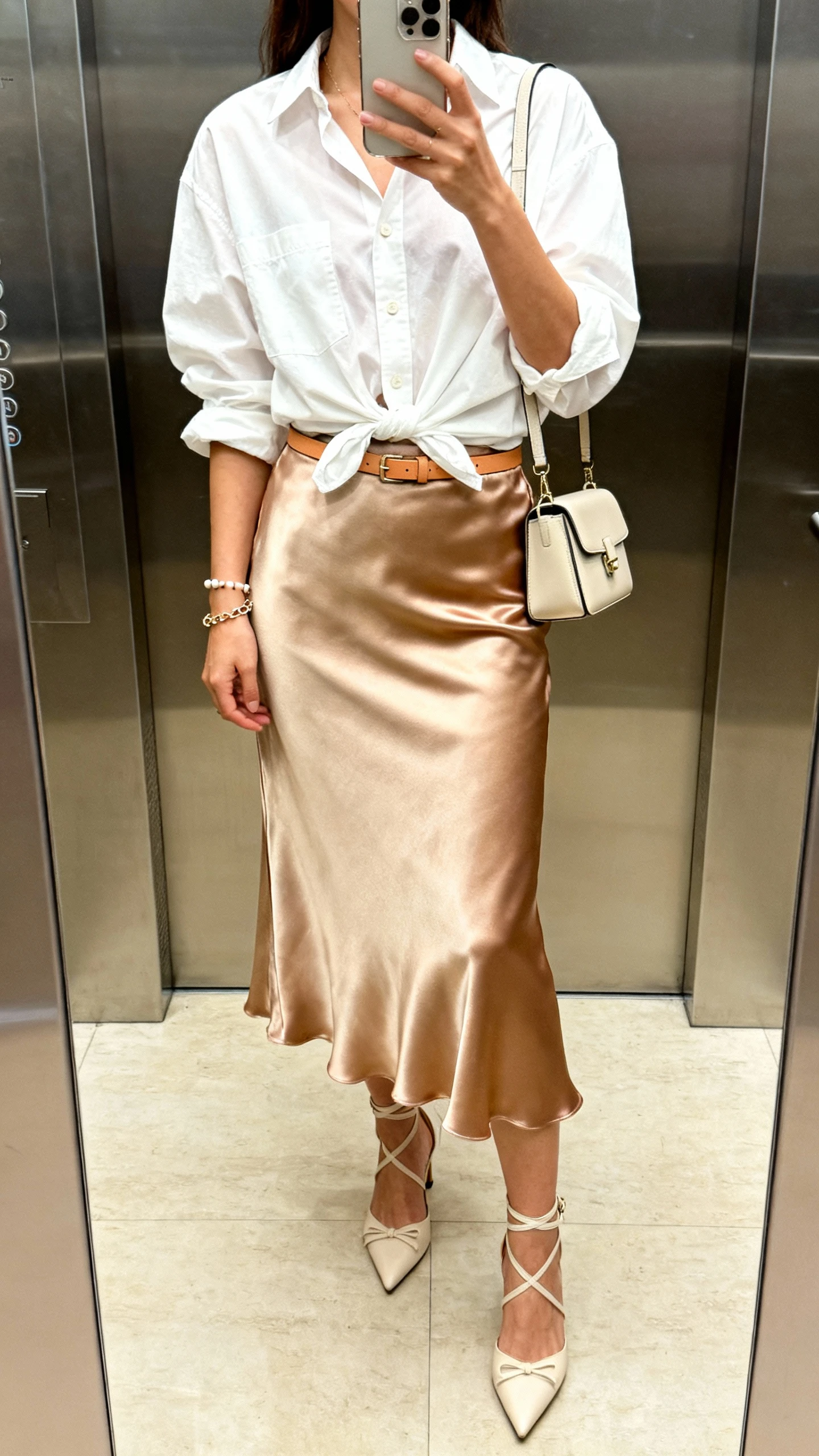 iPhone elevator mirror selfie of a woman in a satin boho midi skirt (bias-cut jewel tone/champagne), slightly oversized crisp white button-down half-tucked, strappy kitten heels, structured mini shoulder bag, delicate chain bracelet with minimalist studs, thin tan leather belt, face not visible, soft indoor lighting, natural iPhone aesthetic.