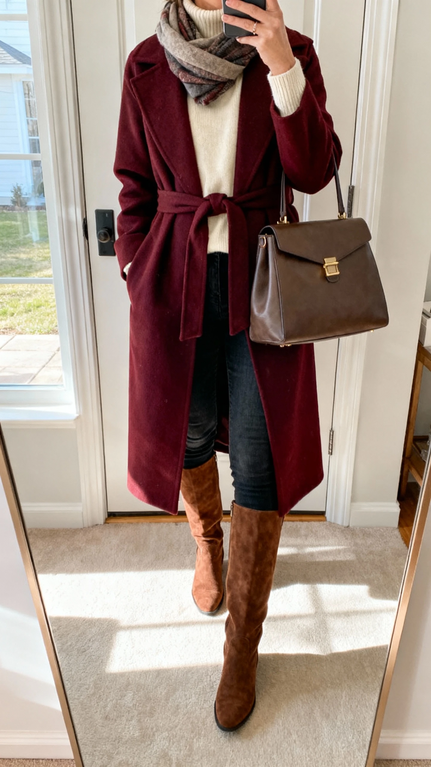 iPhone entryway mirror selfie of a woman wearing a long burgundy wrap coat over a cream turtleneck, dark skinny jeans, suede knee-high boots, scarf, and structured tote, face not visible, morning natural light by the door, natural iPhone photo quality.