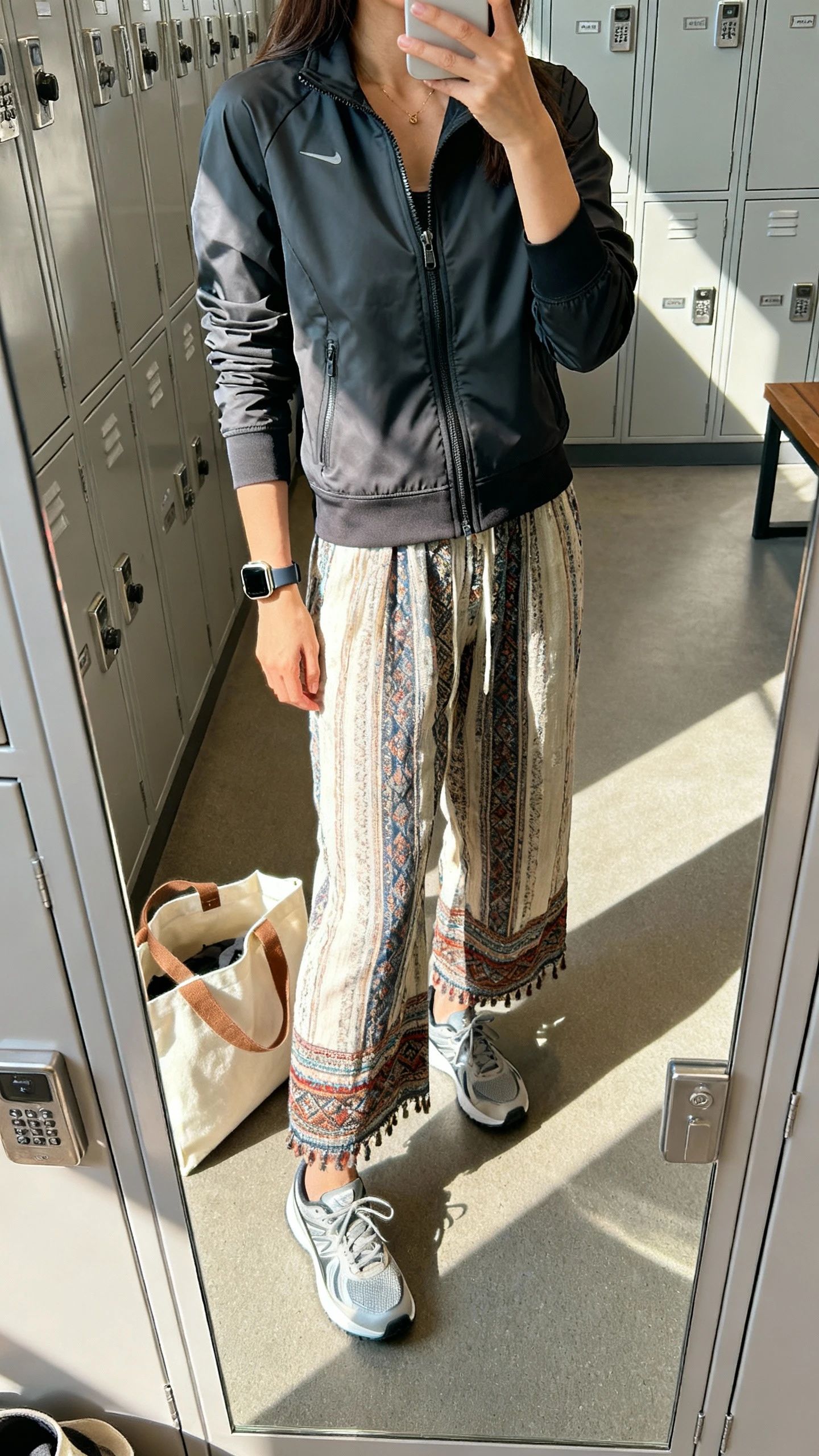 iPhone gym-locker mirror selfie of a woman wearing a sleek zip-up jacket with boho pants and streamlined sneakers, sport watch, tote bag, face not visible, bright morning light, natural iPhone photo quality.