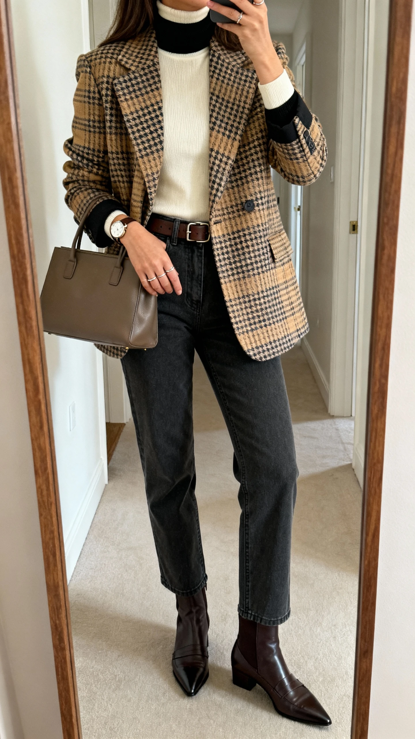 iPhone hallway mirror selfie of a woman in a plaid/houndstooth blazer (tan/charcoal/brown), fitted turtleneck (cream/black), straight-leg jeans or charcoal trousers, loafers or pointed ankle boots, leather belt, minimal watch, delicate rings, structured tote, face not visible, morning indoor light, natural iPhone aesthetic.