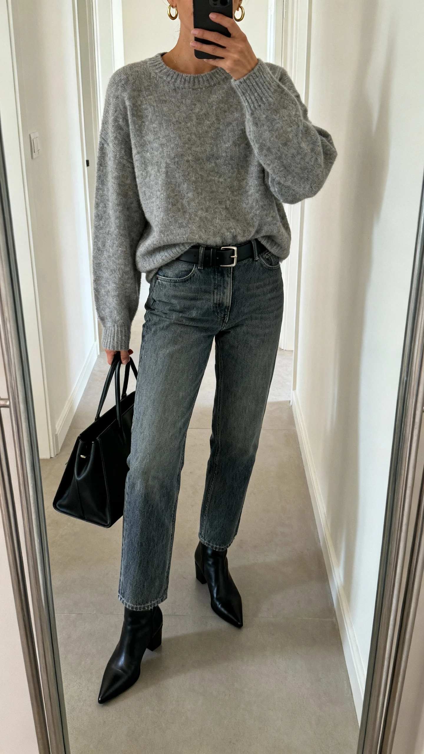 iPhone hallway mirror selfie of a woman in the elevated sweater-and-denim uniform: slouchy heather gray crewneck sweater half-tucked into straight-leg mid-wash jeans, black leather belt, pointed black ankle boots, minimalist gold hoop earrings, structured black tote, face not visible, cropped at neck, soft indoor lighting, natural iPhone quality.