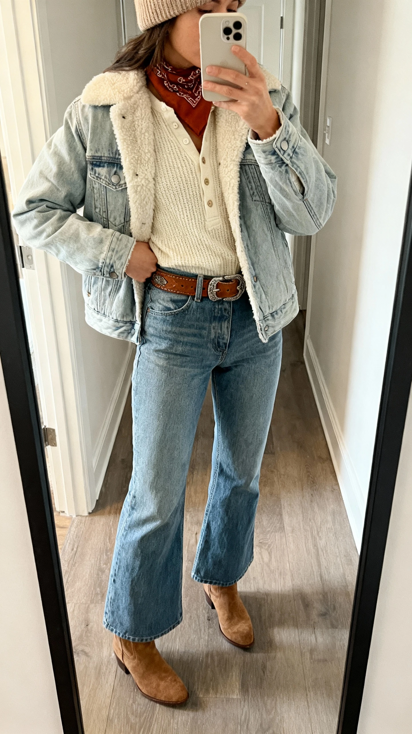 iPhone hallway mirror selfie of a woman wearing a shearling-lined denim jacket (light wash), cream waffle-knit henley, high-rise flare jeans, suede ankle boots, tooled leather belt, rust bandana at neck, wool beanie, face not visible, natural indoor light, casual iPhone photo.