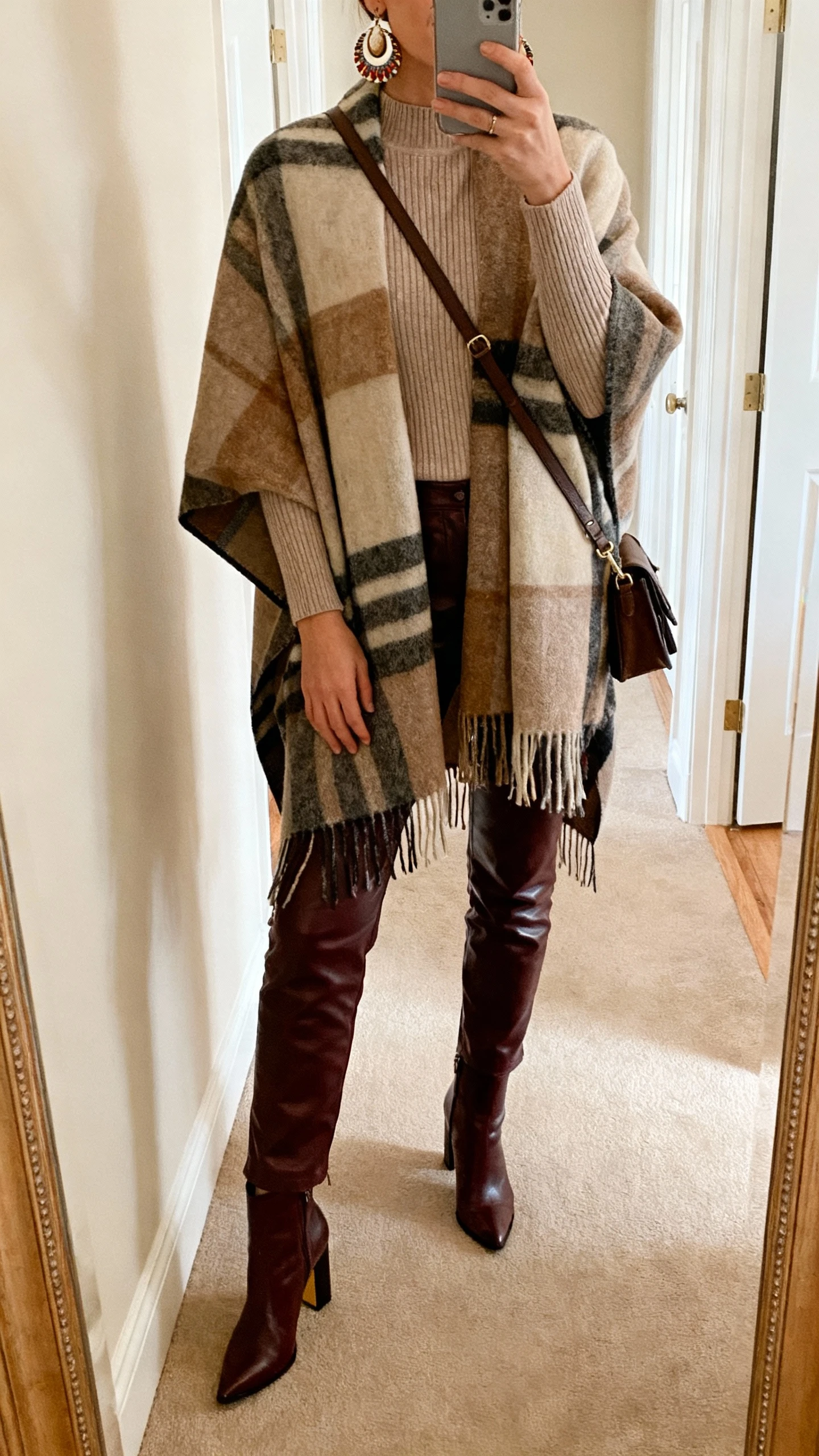 iPhone hallway mirror selfie of a woman wearing a blanket scarf poncho over a fitted knit, leather trousers, heeled ankle boots, and artsy statement earrings with a crossbody bag, face not visible, warm indoor lighting, natural iPhone photo quality.