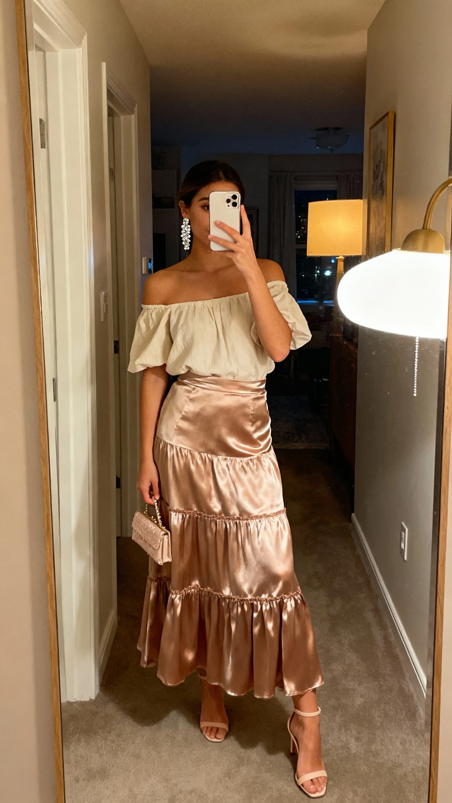 iPhone hallway mirror selfie of a woman wearing a satin long boho skirt with an off-the-shoulder top, delicate heels, dainty clutch, sparkly earrings, face not visible, evening apartment hallway with warm lamp light, natural iPhone aesthetic