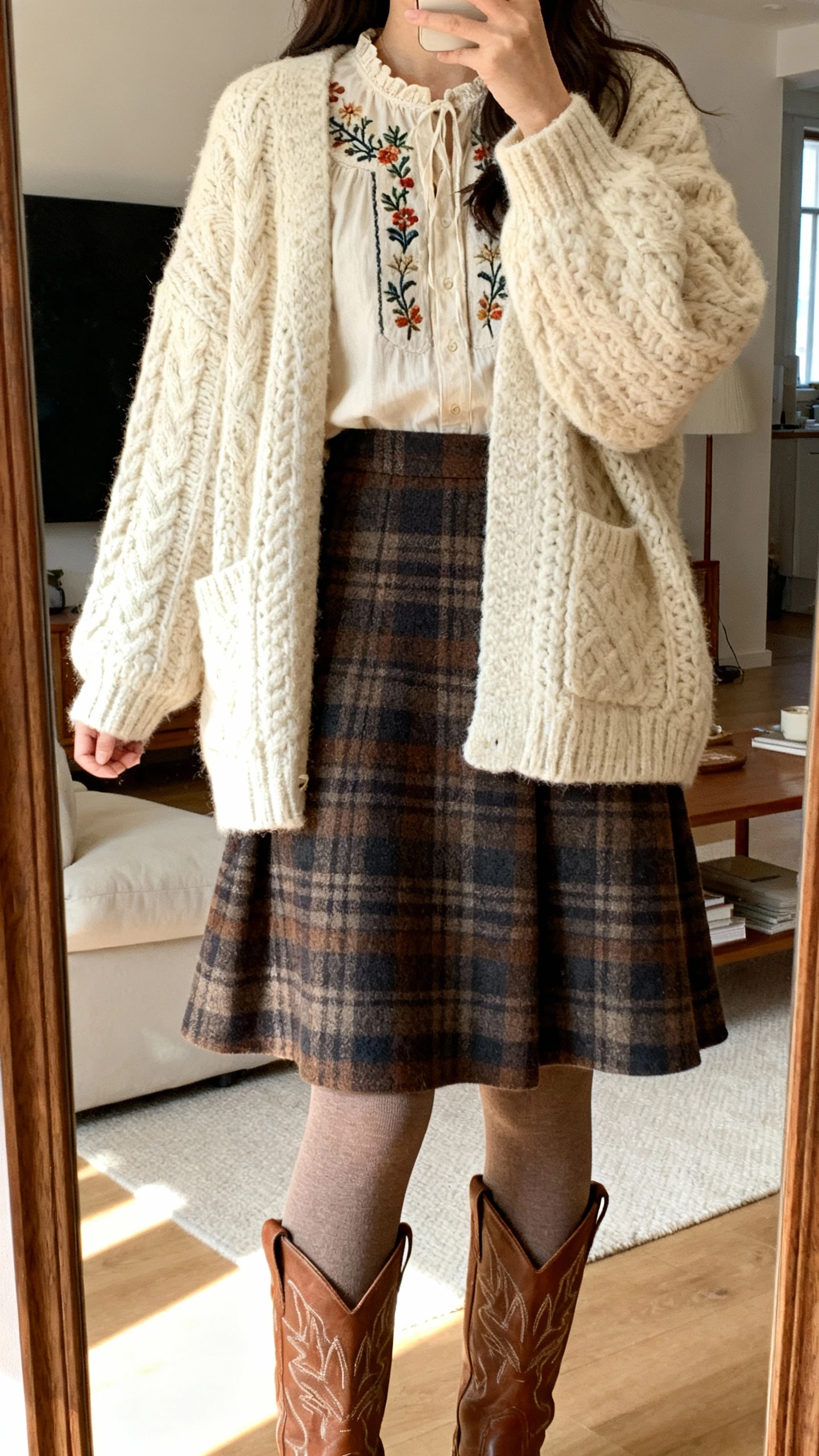 iPhone mirror selfie cropped at shoulders showing a woman wearing an oversized cream chunky cardigan over an embroidered blouse, plaid A-line wool skirt, thermal tights, Western knee-high boots, face not visible, cozy living room mirror with soft indoor light, natural iPhone aesthetic.