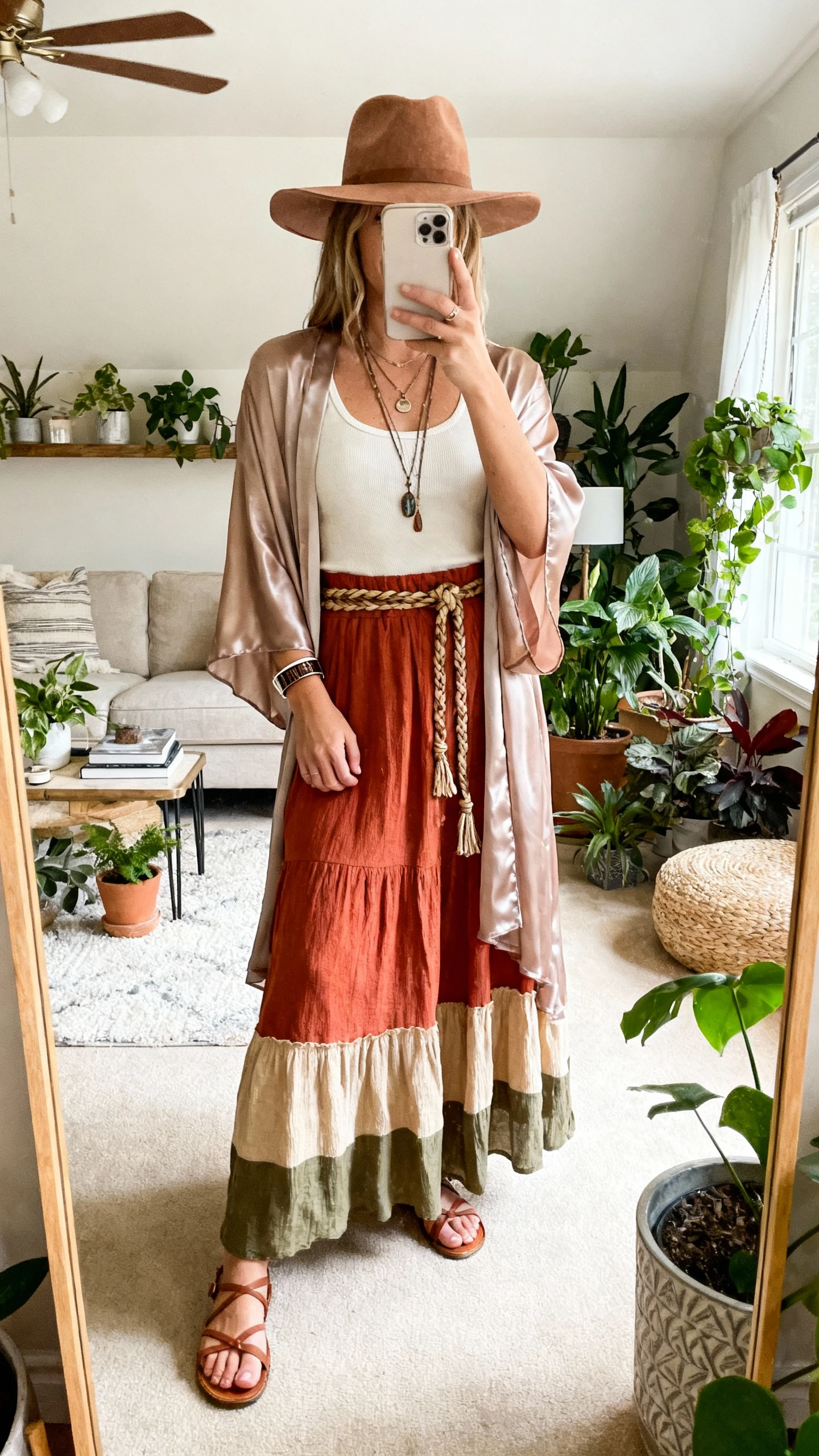 iPhone mirror selfie in a plant-filled living room of a woman in a flowy boho maxi skirt (terracotta/sand/olive), simple cream tank, silky chiffon kimono, strappy flat sandals, braided cinch belt, layered pendant necklaces with a cuff bracelet, wide-brim felt hat, face not visible, soft morning light, natural iPhone photo quality.