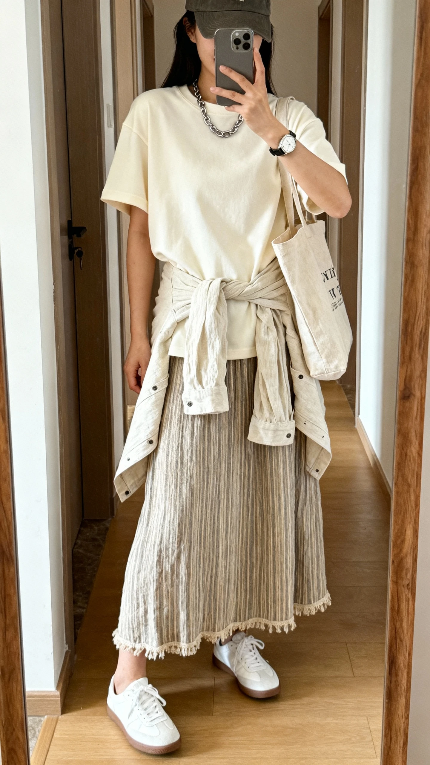 iPhone mirror selfie in hallway of a woman in a linen boho skirt (solid or subtle stripe), oversized cream crewneck tee, white sneakers, canvas tote, baseball cap, simple chain necklace and watch, lightweight linen shacket tied around waist, face not visible, bright natural daylight, iPhone photo quality.