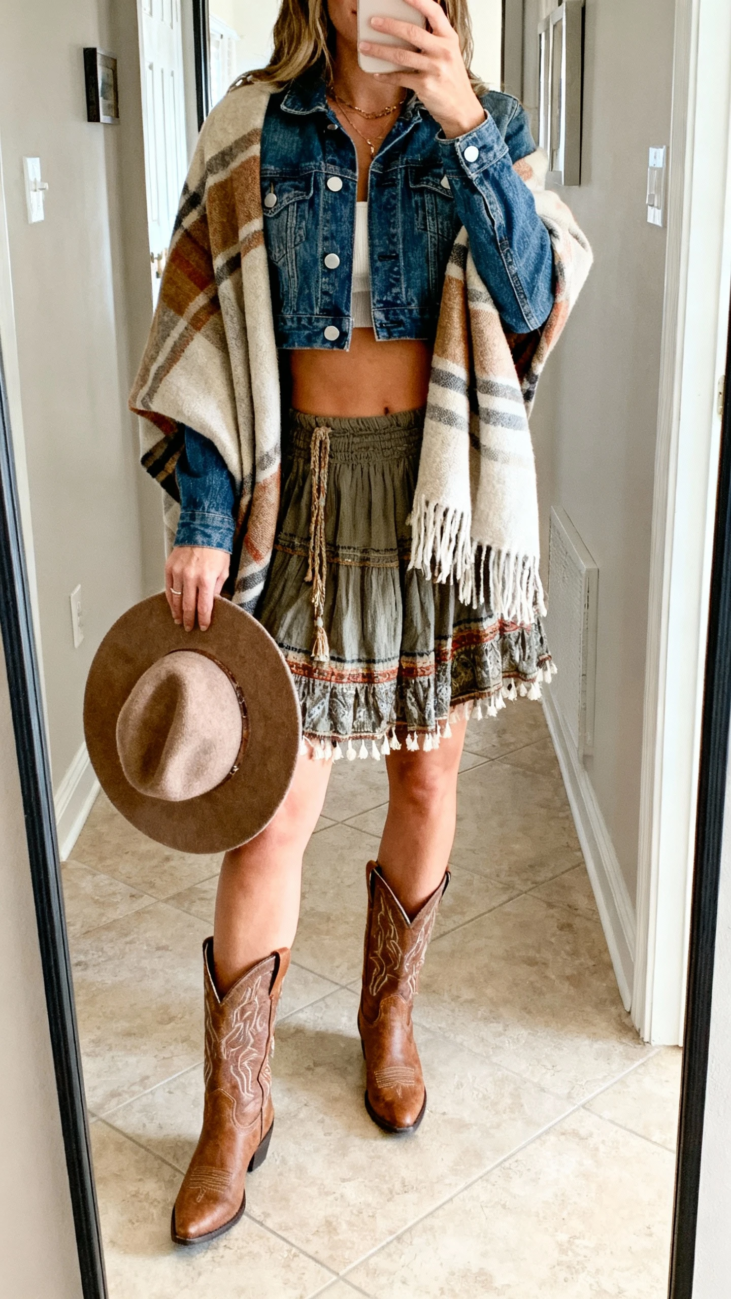 iPhone mirror selfie of a woman in a cropped denim jacket layered over a boho skirt with a blanket scarf wrapped around the shoulders, western ankle boots, and a felt hat held in hand, face not visible, entryway full-length mirror, bright natural daylight, casual iPhone photo.