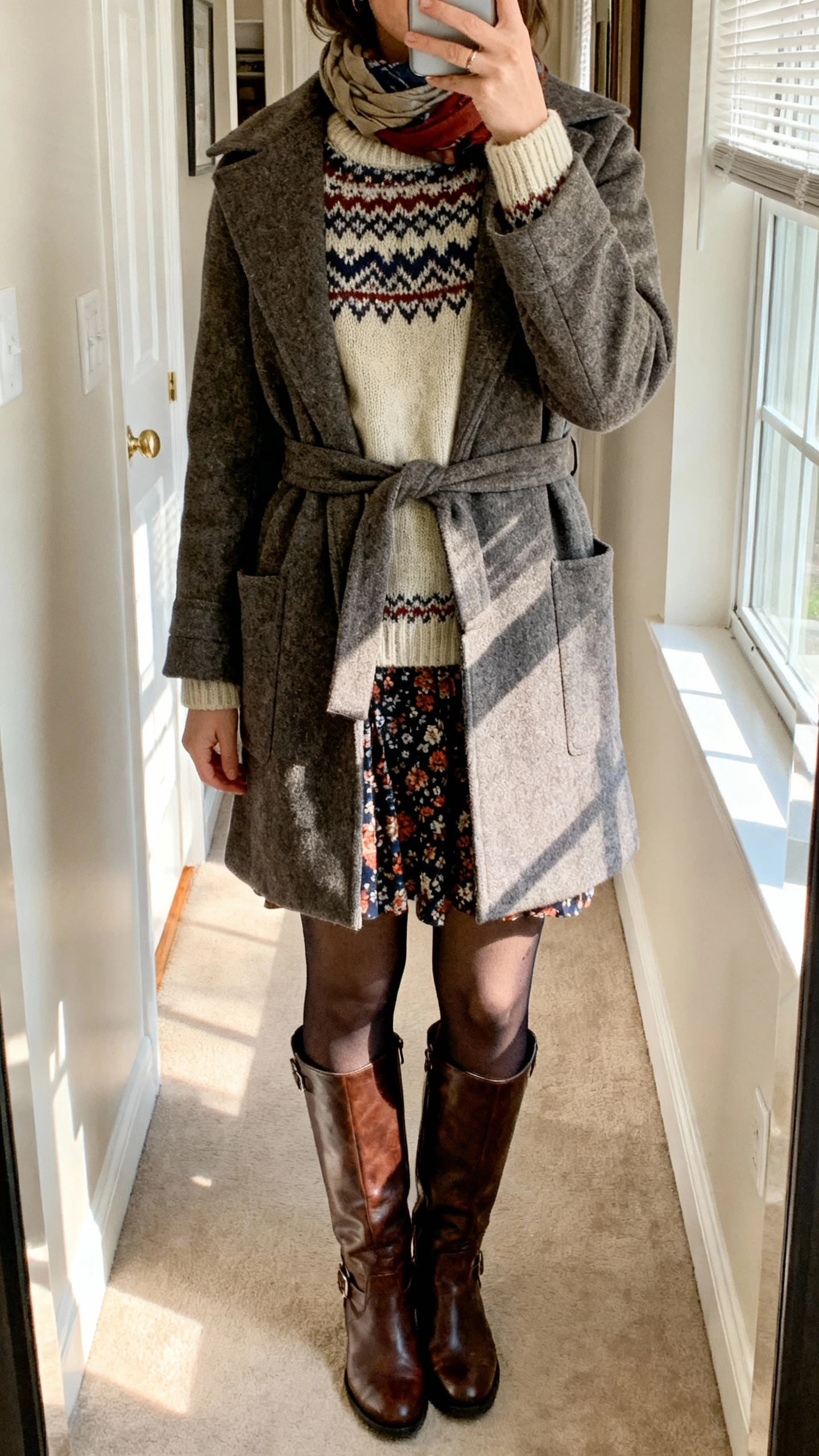 iPhone mirror selfie of a woman in a Fair Isle knit sweater under a belted wool wrap coat paired with a printed skirt, opaque tights, leather knee boots, and a scarf peeking out, face not visible, apartment hallway mirror, afternoon window light, natural iPhone aesthetic.