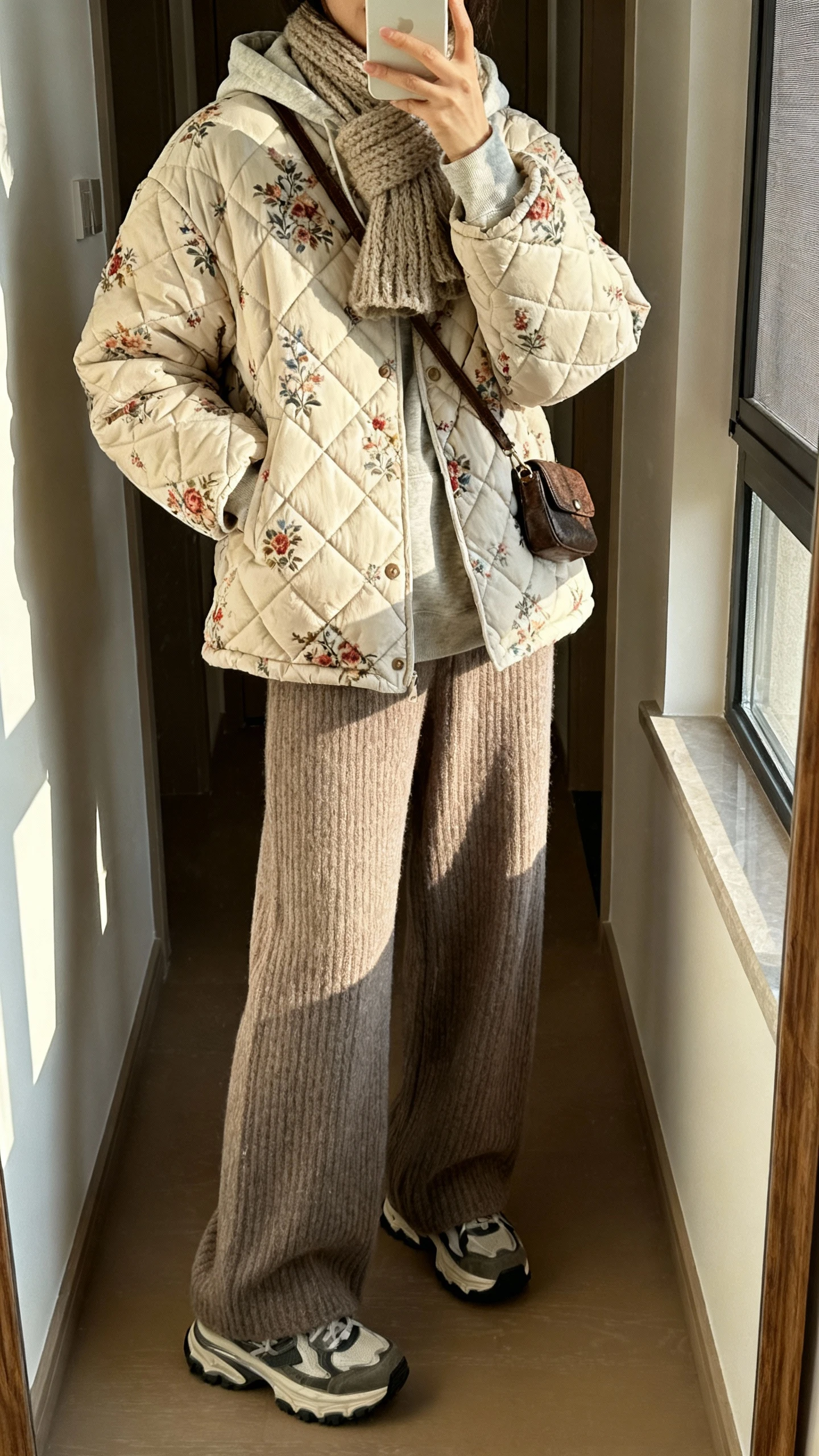 iPhone mirror selfie of a woman in a quilted boho puffer with floral quilting, cozy hoodie, wide-leg knit pants, chunky sneakers, knit scarf, and small crossbody, face not visible, hallway mirror with morning window light, natural iPhone photo quality.