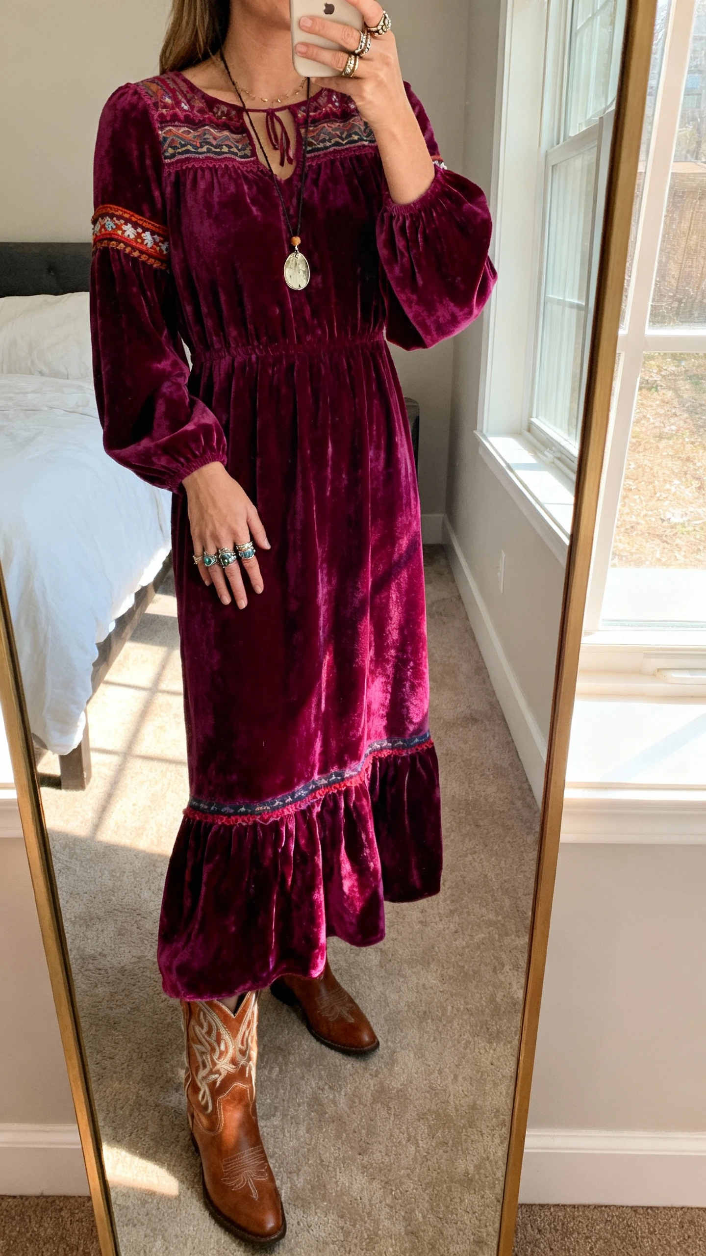 iPhone mirror selfie of a woman in a rich jewel-tone velvet maxi dress with a folk-tale twist, western boots, layered boho rings and a pendant, face not visible, cropped at neck, bedroom full-length mirror, warm window light, natural iPhone photo quality.