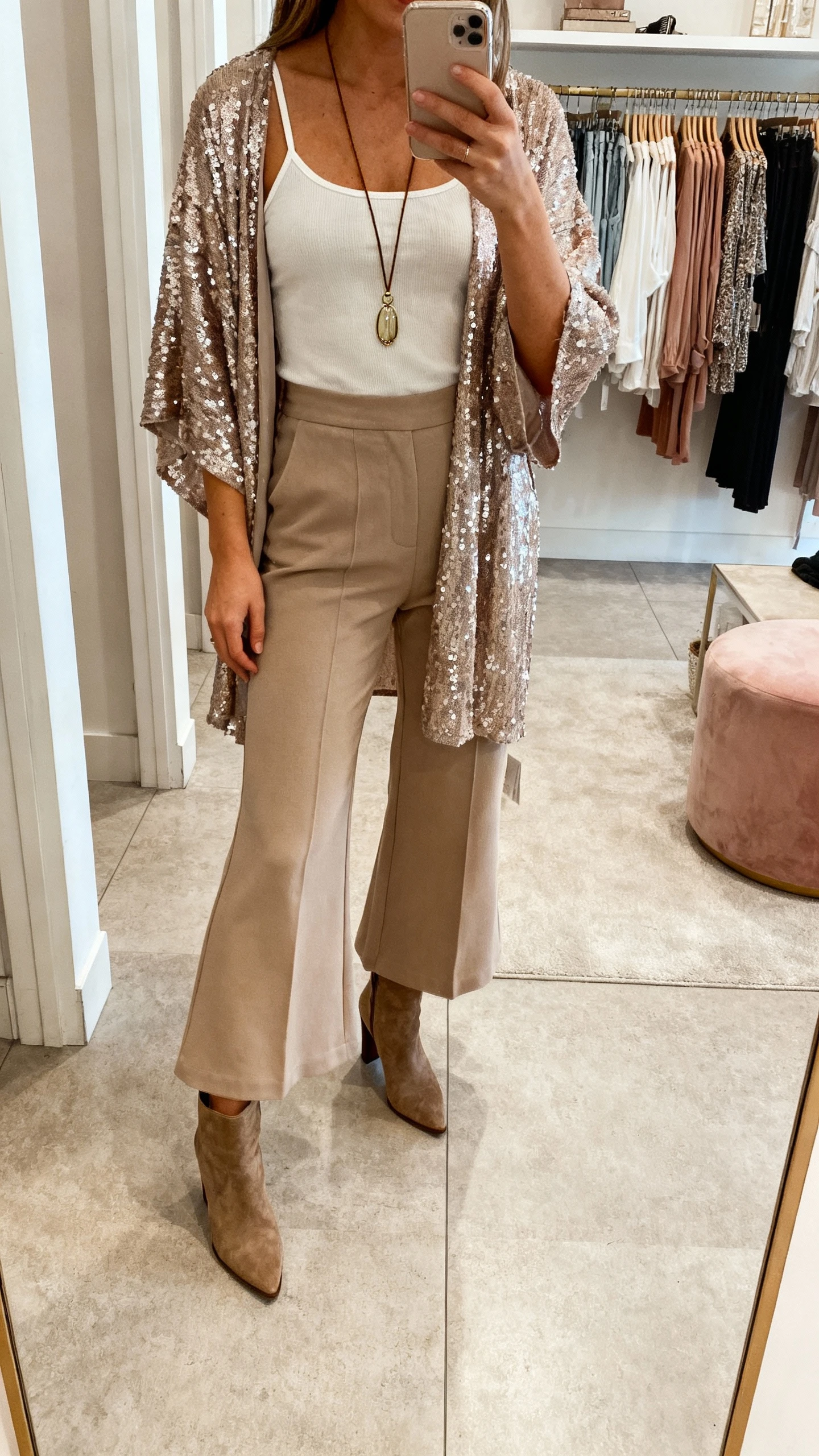 iPhone mirror selfie of a woman in a sequin kimono layered over high-waisted flares with a simple camisole, heeled boots, long pendant necklace, face not visible, boutique dressing room mirror, indoor lighting, natural iPhone photo quality.