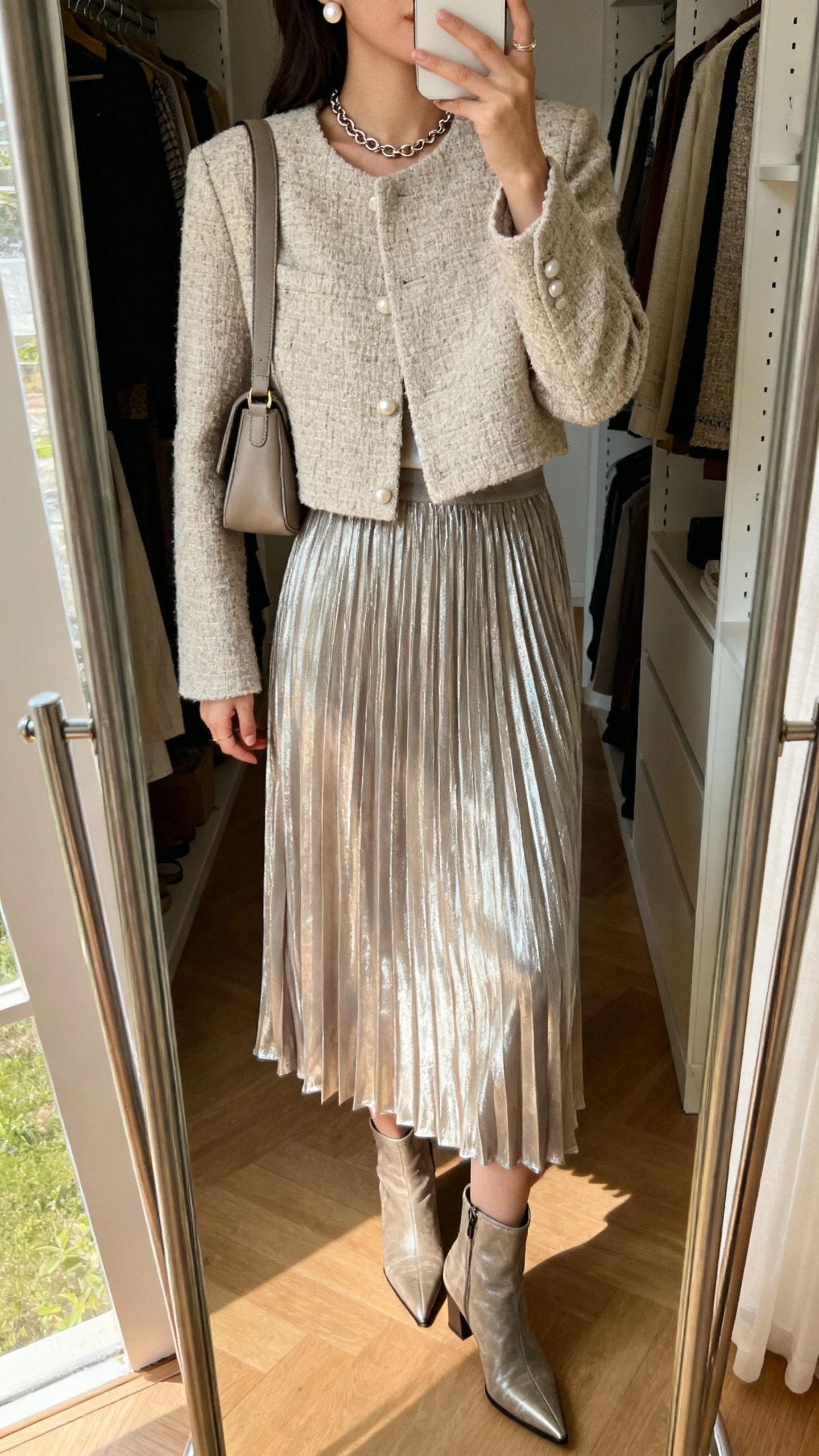 iPhone mirror selfie of a woman in a softly metallic pleated midi skirt with a neutral tweed cropped jacket, pointed ankle boots, a short chain necklace, pearl studs, and a medium shoulder bag, face not visible, full-length closet mirror, afternoon natural light.
